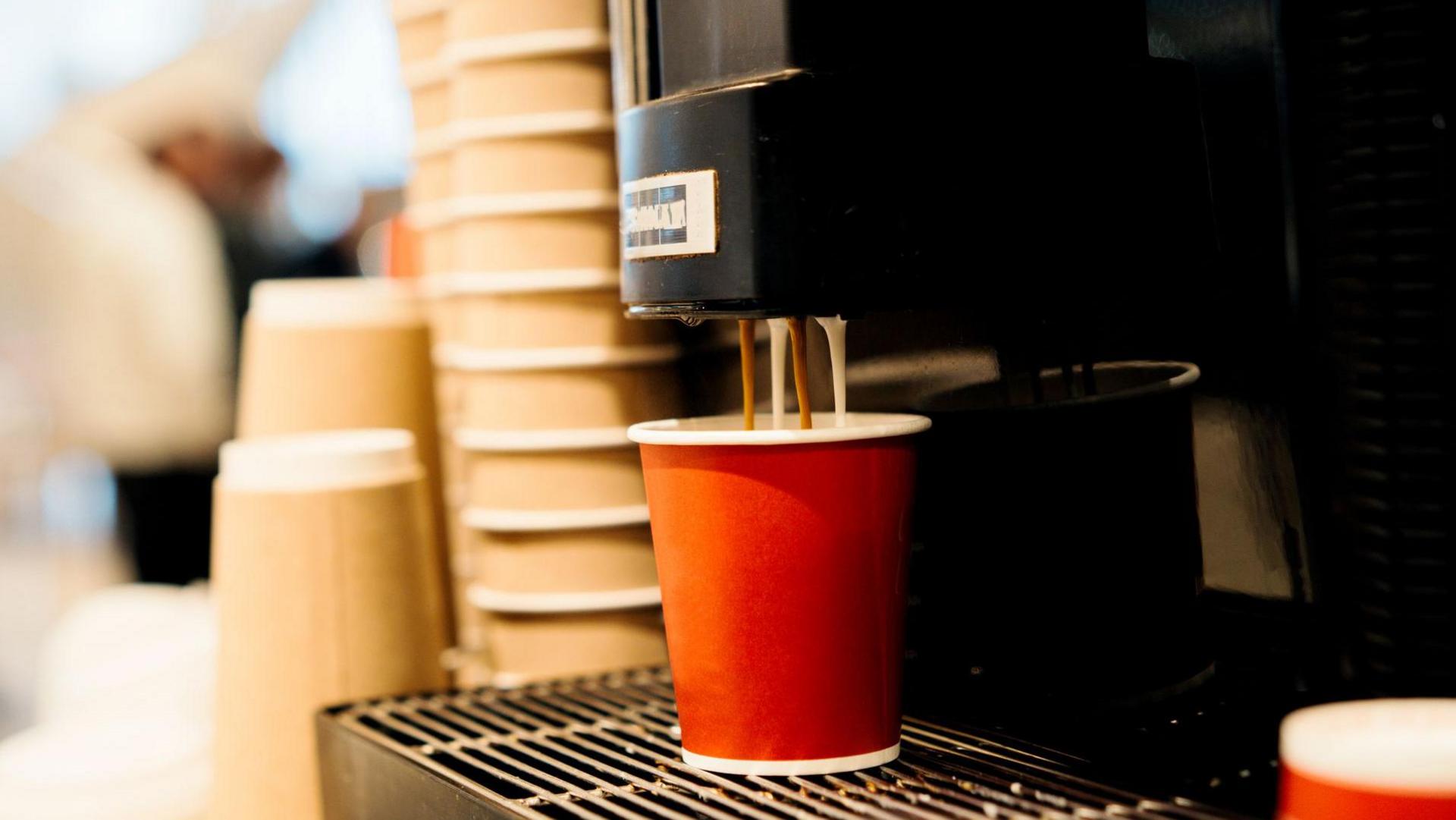 A coffee machine dispensing milk and coffee into an orange paper cup. There is a tower of brown papers cups in the background.