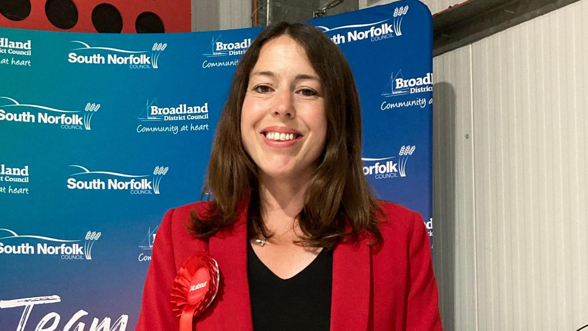 Labour MP Alice Macdonald wearing a red jacket and red Labour Party rosette. She is smiling and standing in front of a screen advertising South Norfolk and Broadland councils.
