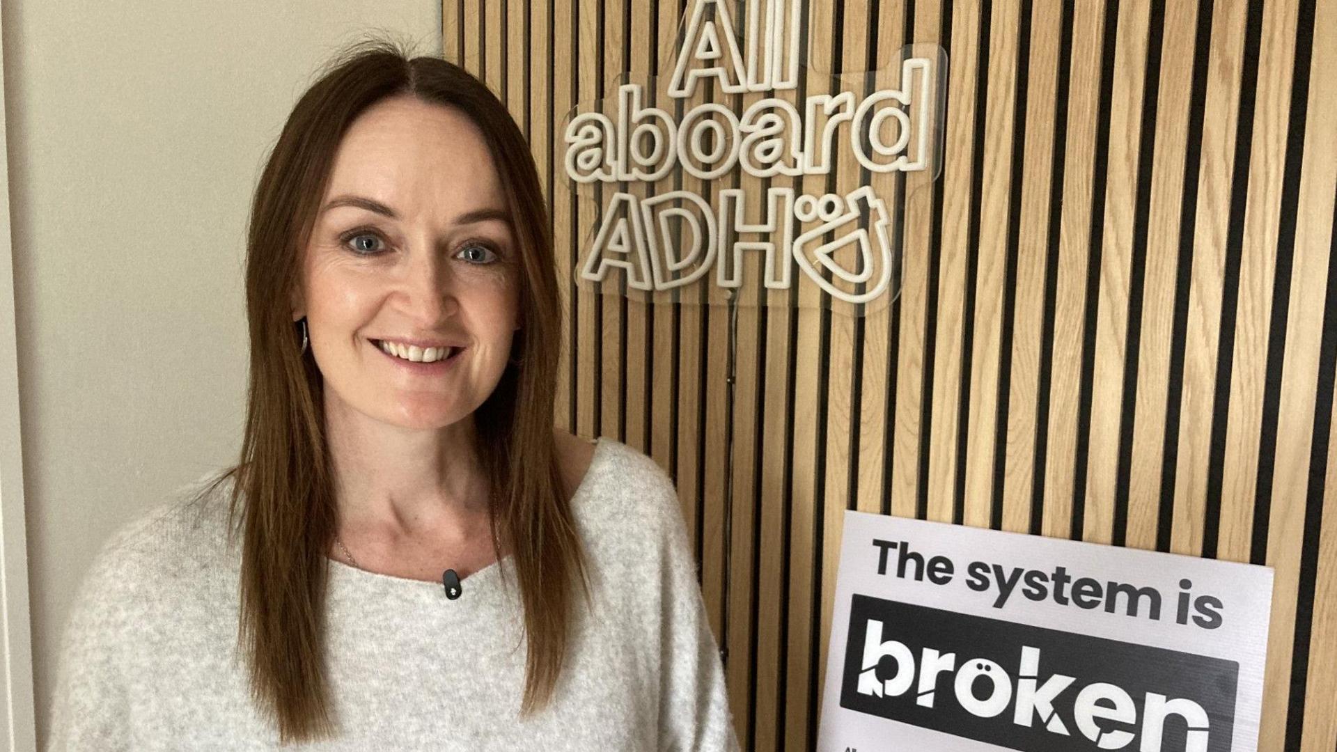 A woman with long brown hair in a light jumper stands in front of wood panelling. One sign on the wall behind her reads "All Aboard ADHD." A sign below it reads "The system is broken - not our kids"