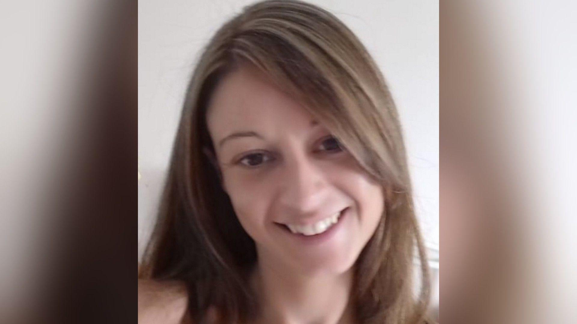 A portrait image of a woman with long brown hair smiling while in front of a white wall.