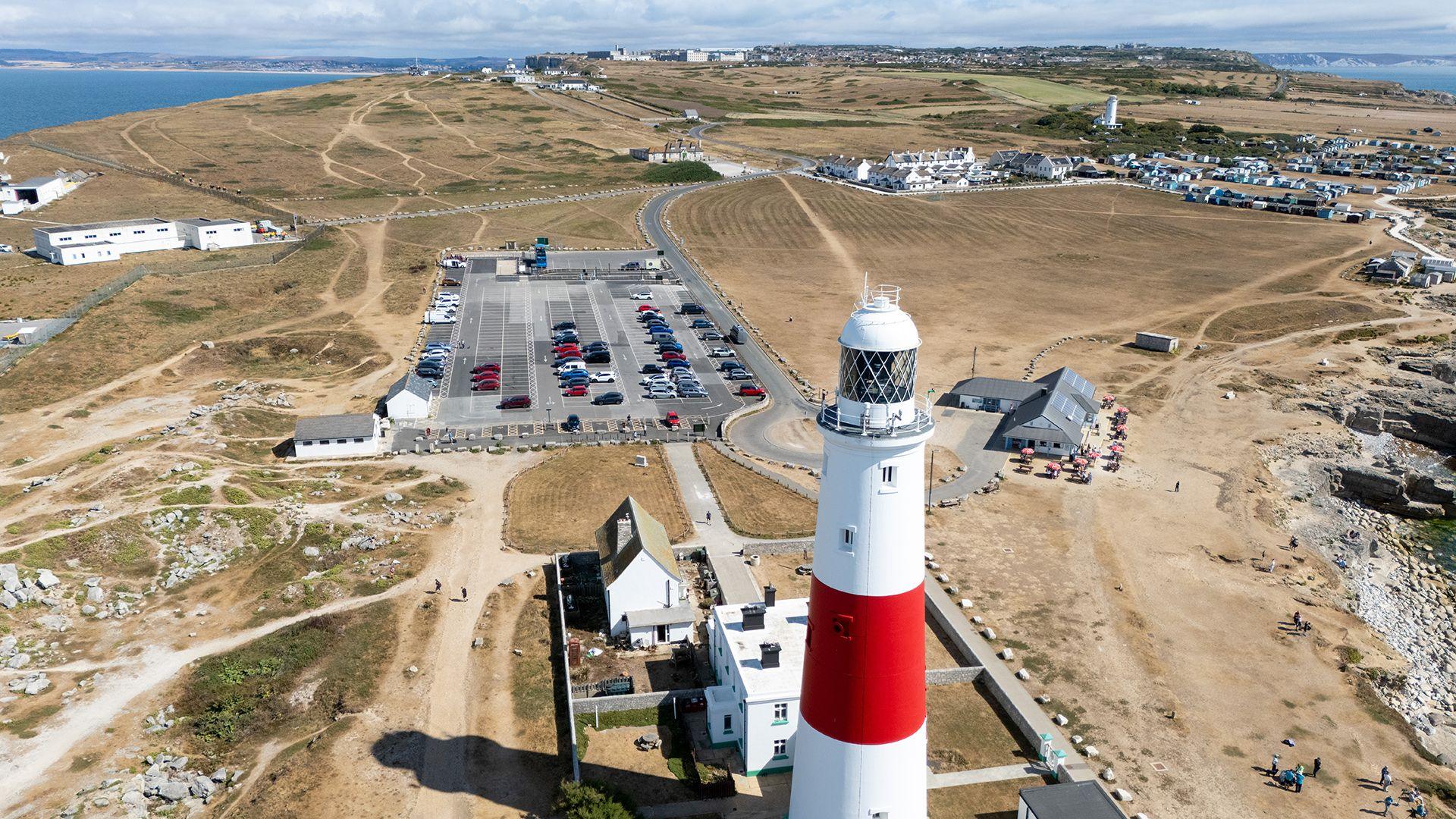 An aerial view shows Portland lighthouse next to a car park and a wide view of brown grass and vegetation, with the sea and a cloudy blue sky in the background, taken on Thursday.