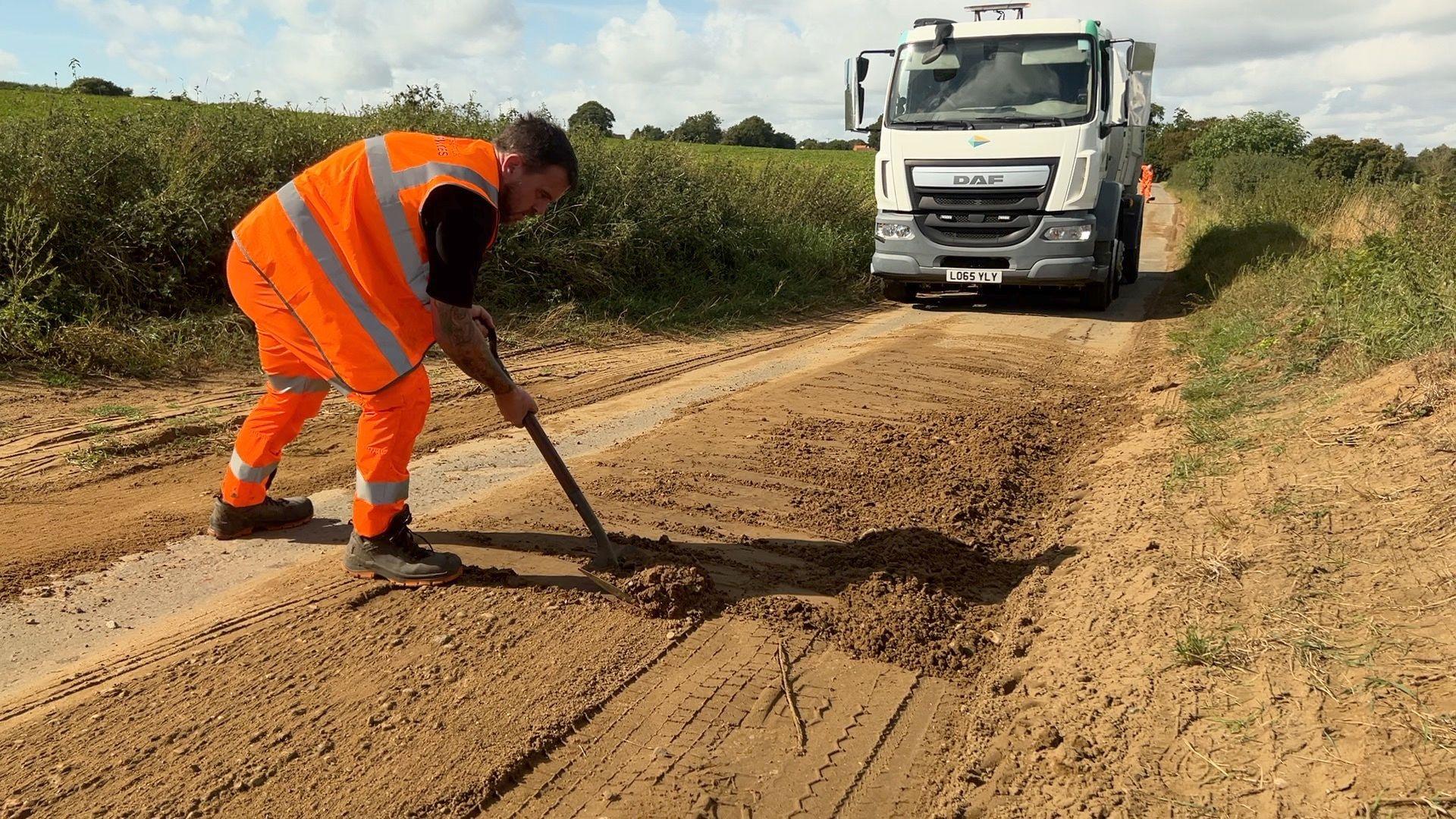 A highways worker wearing an orange high vis uniform rakes mud from a road to clear it. A HGV lorry is parked in the distance.