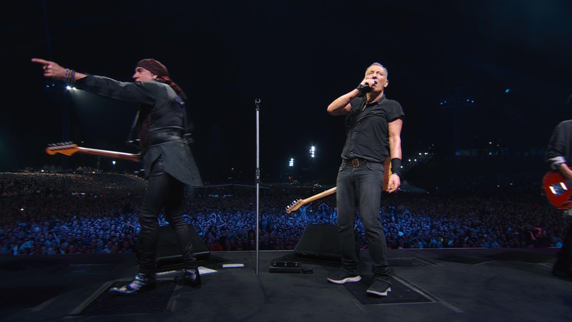 Bruce Springsteen (right) and Steve Van Zandt on stage during his world tour, the pair are both dressed in black and holding guitars. Van Zandt is looking out on the crowd and pointing while Springsteen is looking towards the back of the stage.