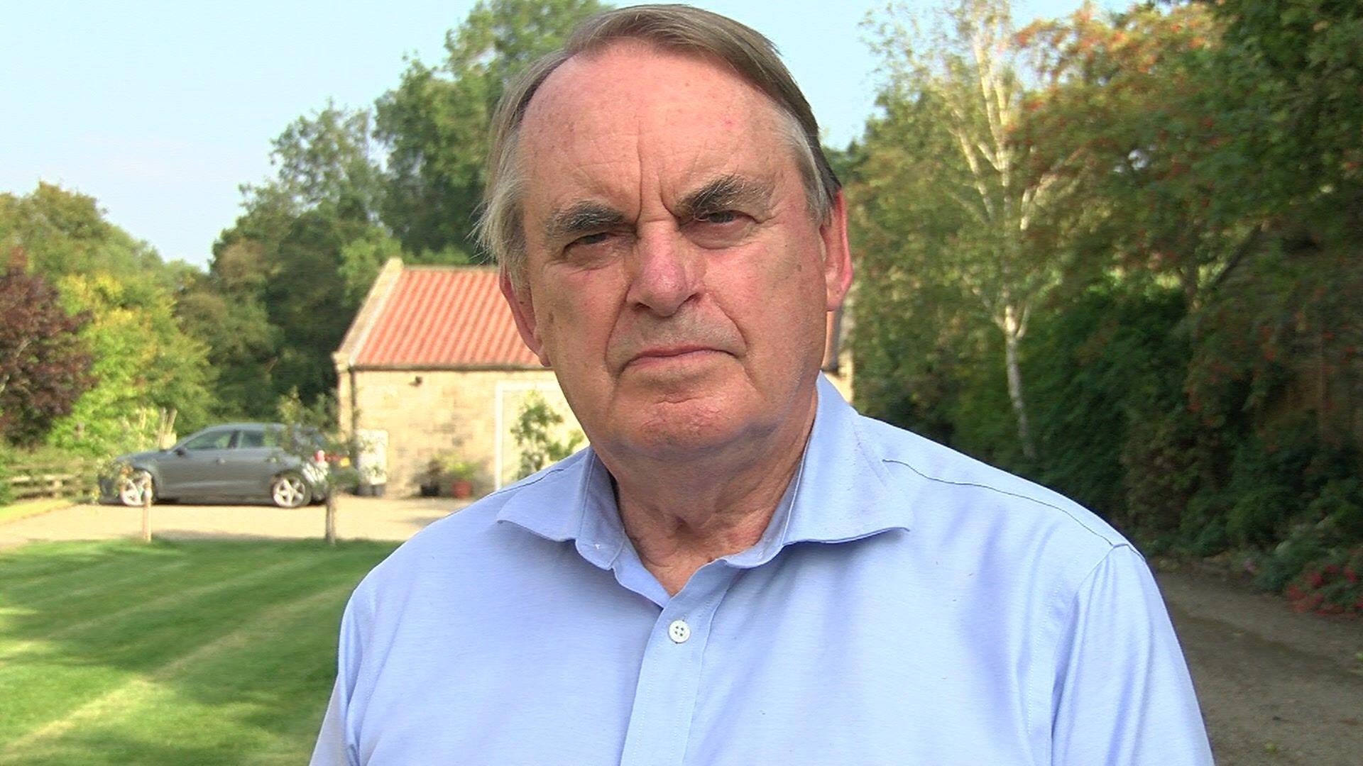 Head-and-shoulders shot of Lord Kirkhope who has receding mousey-brown hair, with grey sections by his temples, and is wearing a light blue shirt. He is standing in a lawned area with trees and shrubs on the right. There is a stone outbuilding in the background with a grey car parked next to it.