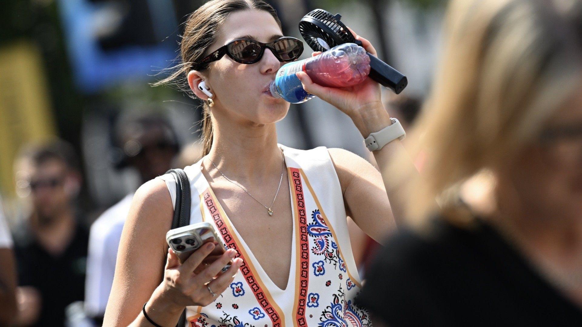 A pistillate   drinks from a bottle, holding a portable fan, with a blurred representation   of radical   successful  the background