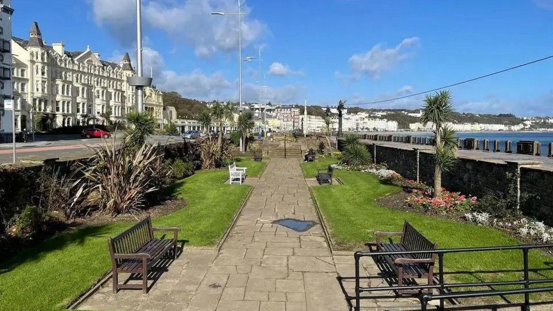 A view from the steps into one of Douglas Promenade's sunken gardens. There is a slabbed path down the middle with benches on either side and some grass, trees, and shrubs lining the sides.