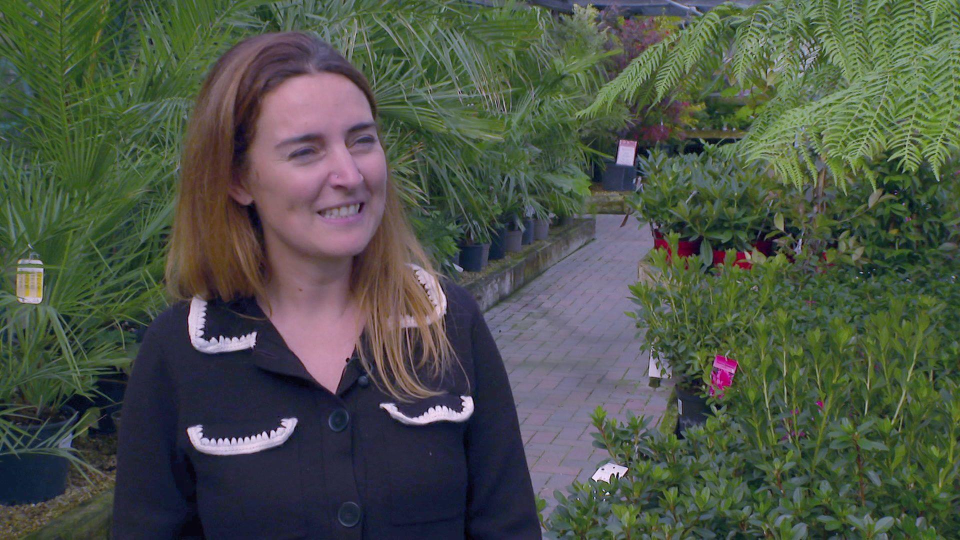 Amy Stewart smiles during a TV interview. She has long fair hair and is wearing a black blouse with white lace trim on the collar and breast pockets. She is standing among large palms and ferns in the plant sales area of the garden centre.