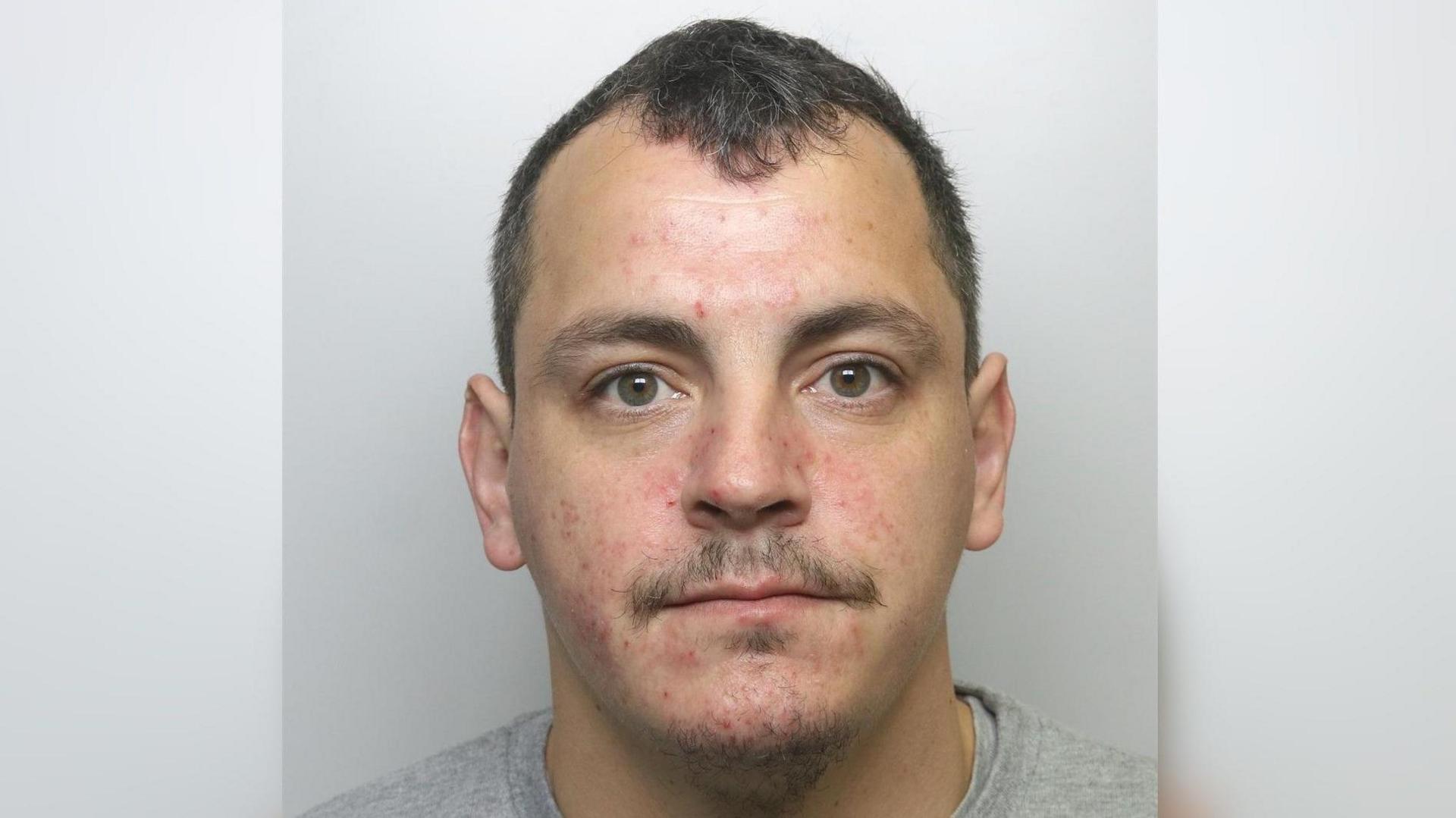 A police headshot of a man with short dark hair wearing a grey T shirt.