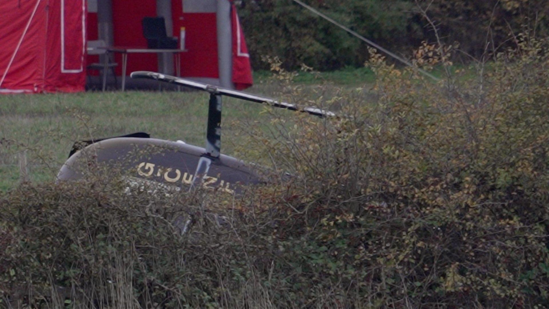 A black helicopter on its side in a field, partly concealed behind some bushes.