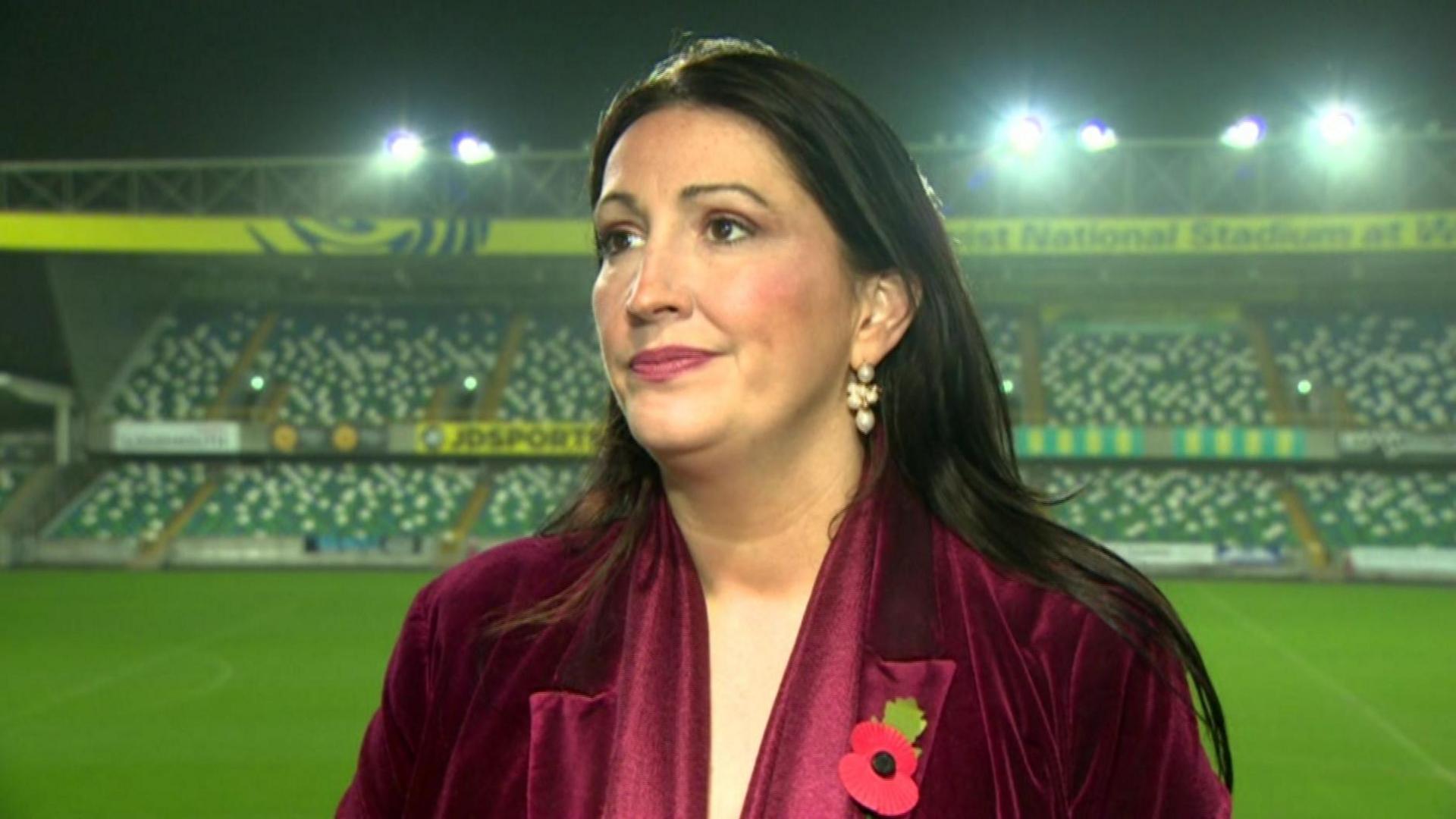 A woman wearing a maroon top and wearing a poppy - standing in a football stadium with the pitch and stand in view behind him.