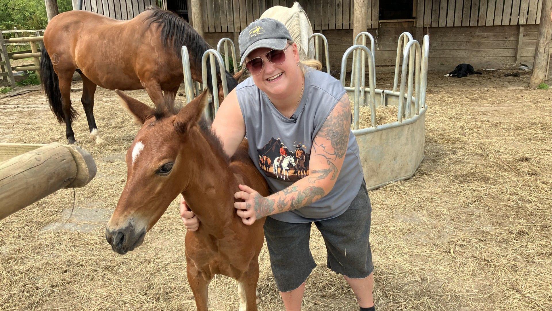 A woman, wearing sunglasses and a cap, smiles at the camera. She has her arms around a brown foal. There are two horses feeding from a trough in the background, with a barn behind them.