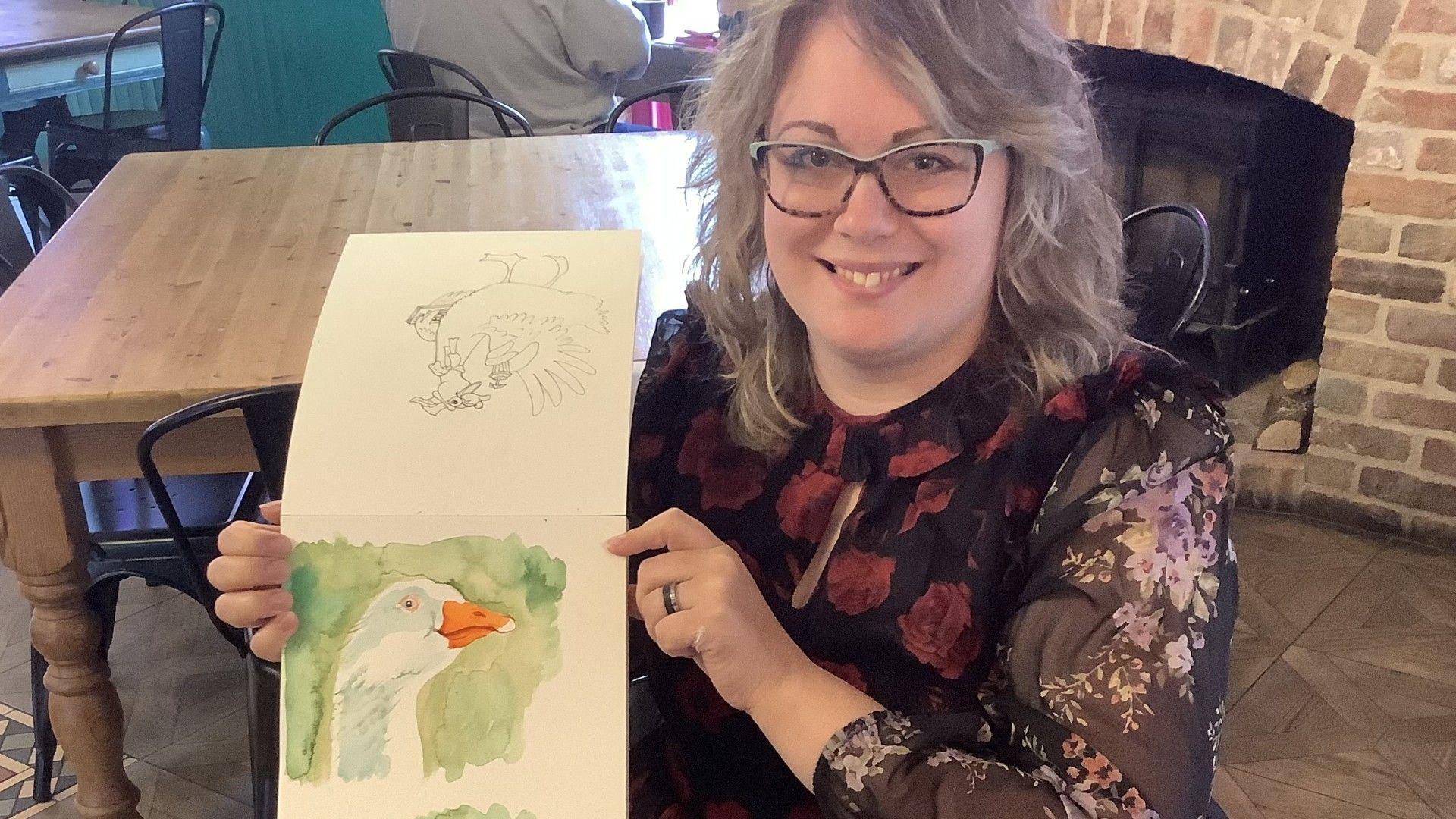 Victoria Jane Clark has mousy, shoulder-length hair and tortoiseshell-framed glasses. She is wearing a floral dress and is holding a watercolour painting of a white goose with an orange bill. Behind her are some tables and chairs in a pub, with an unlit fireplace in the background.