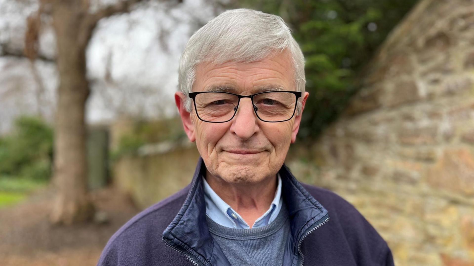 Allan is wearing glasses and is smiling. He is wearing a blue fleece over a lighter blue jumper and shirt. He is standing in a walled garden.