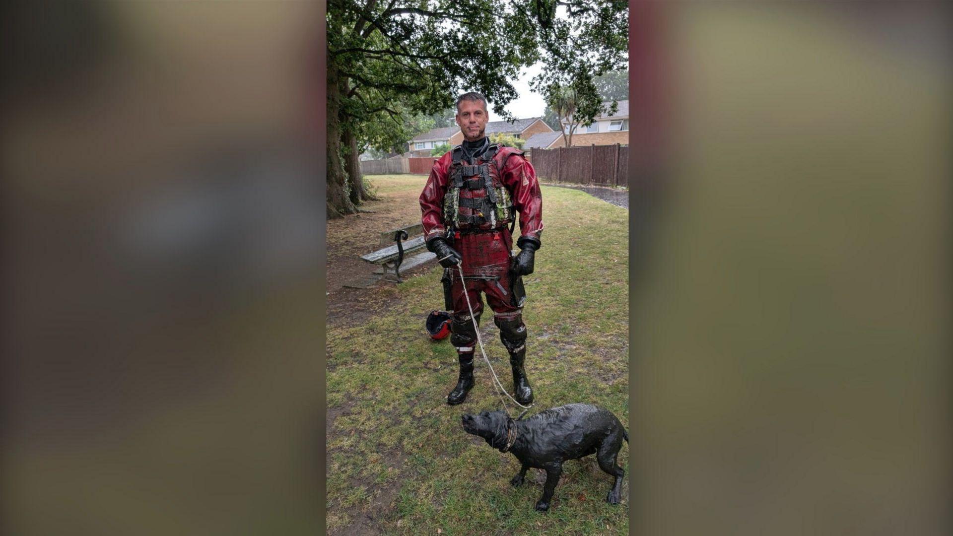 The firefighter is standing, looking at the camera and holding the lead, while Bailey stands in front of him, mid-shake. The firefighter is wearing the same red uniform, with black gloves and boots on.