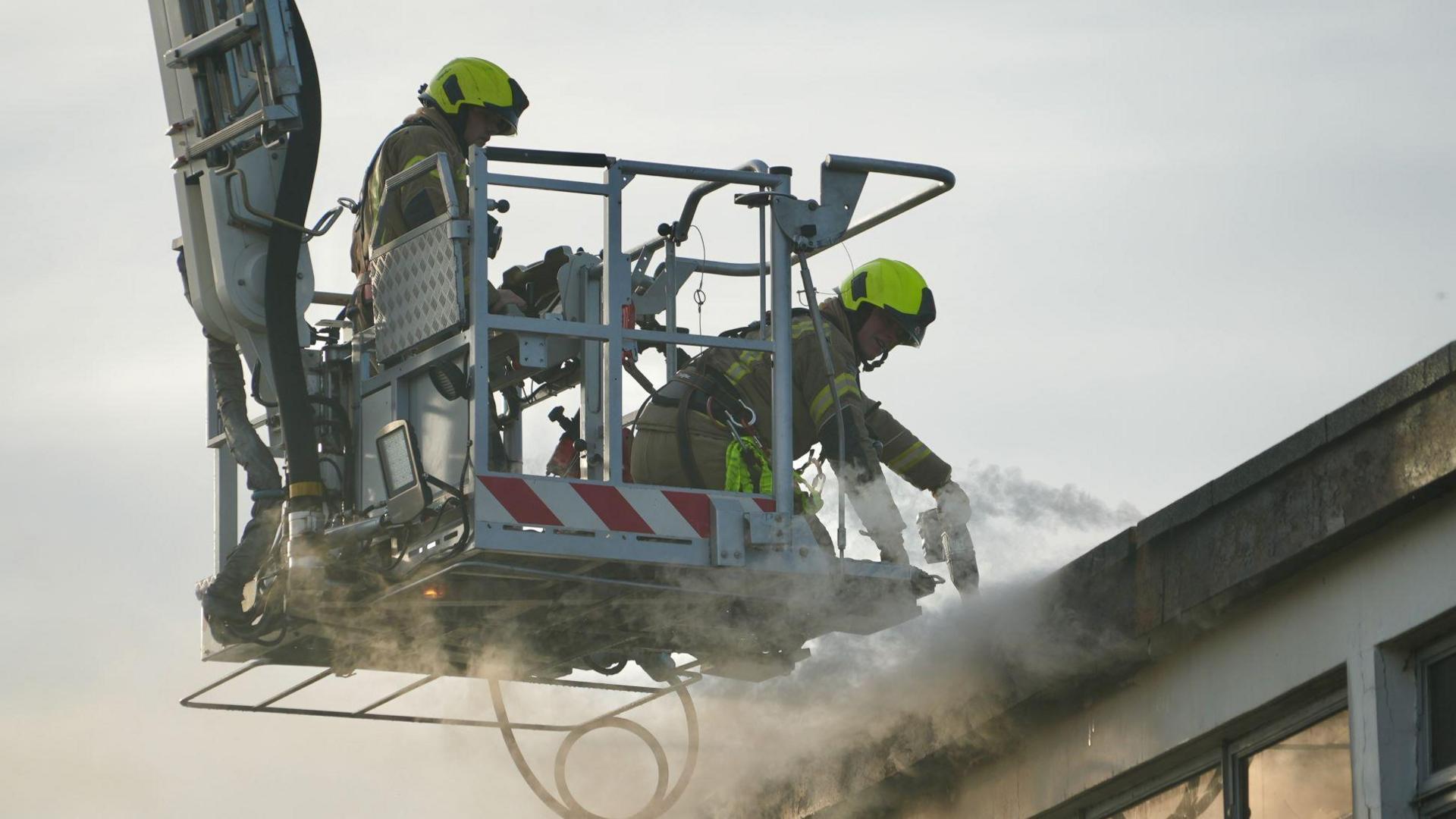 Two firefighters on an aerial ladder platform putting out the fire. There is smoke coming out of the top of the building.