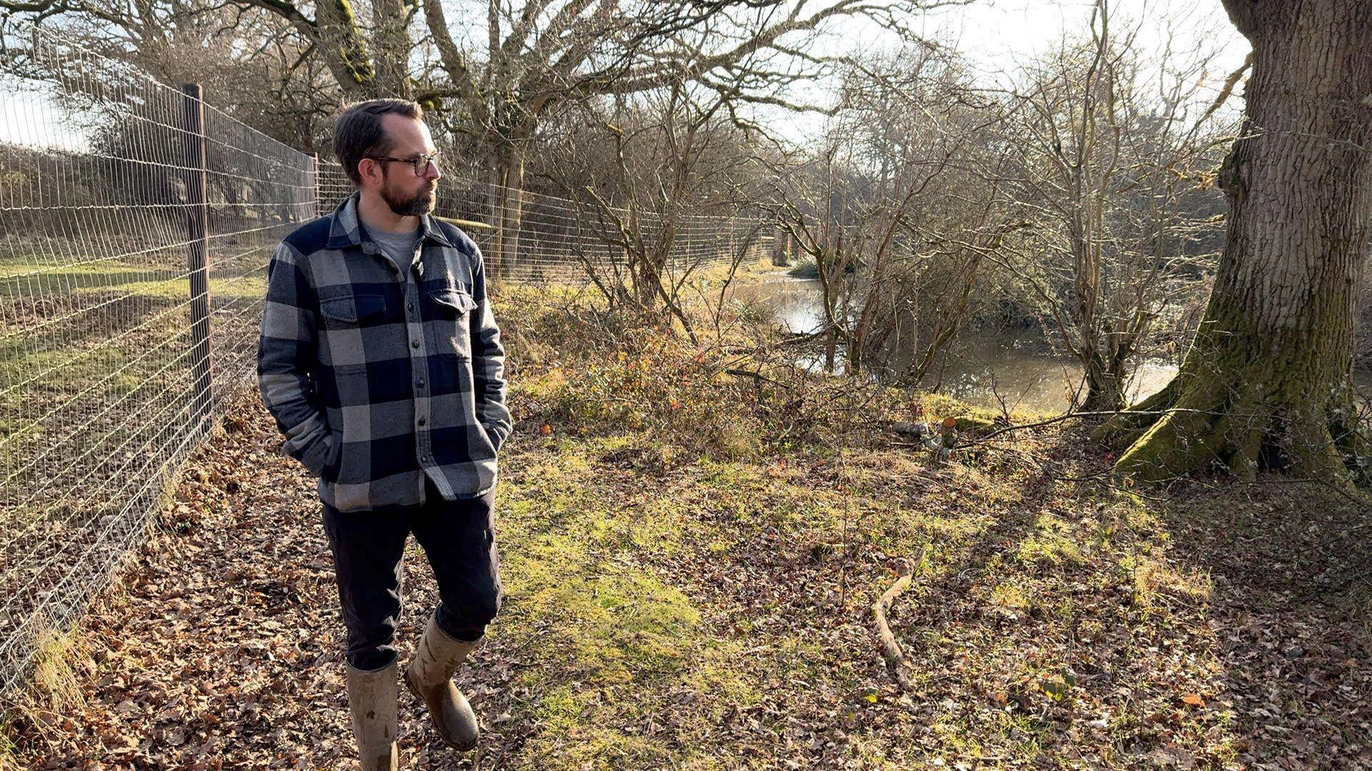 Matt, a bearded man in a checked shirt with glasses walks beside the beaver pool.