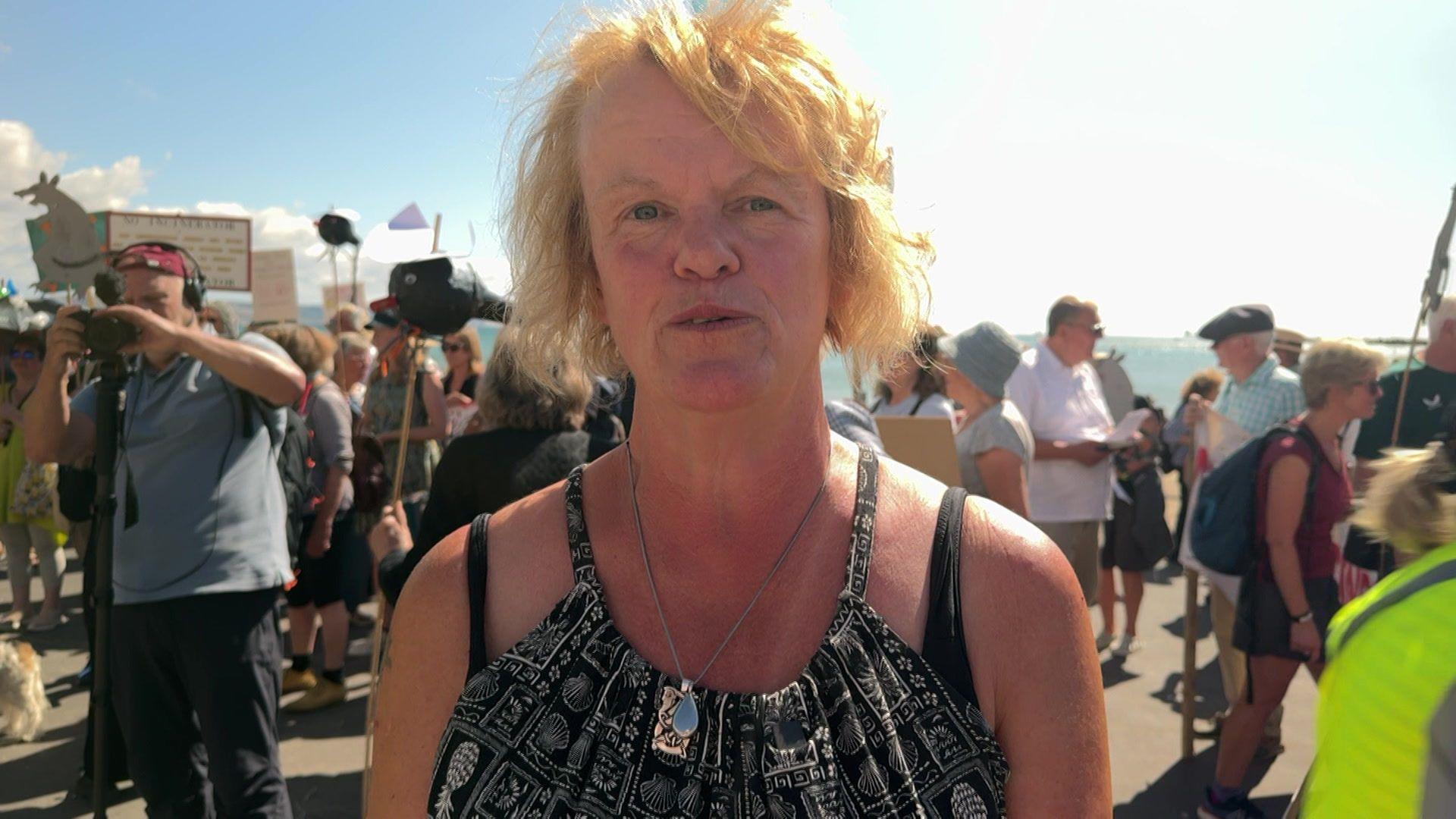 Jill Johnstone looking at the camera, has blonde hair, blue eyes and is wearing a strappy top. There are campaigners in the background.
