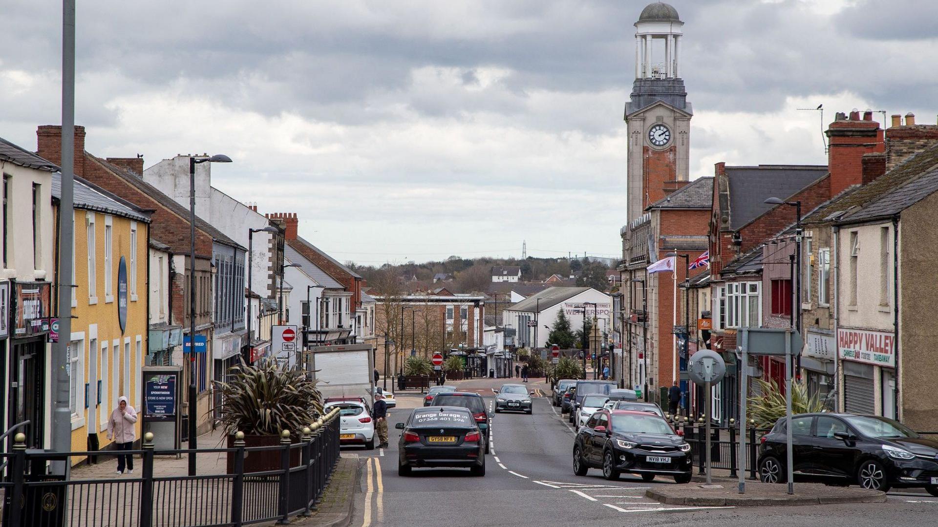 Spennymoor town centre. Cars driver down the high street. A number of closed shops are visible to the right. A clock tower can be seen at the far end of the road.