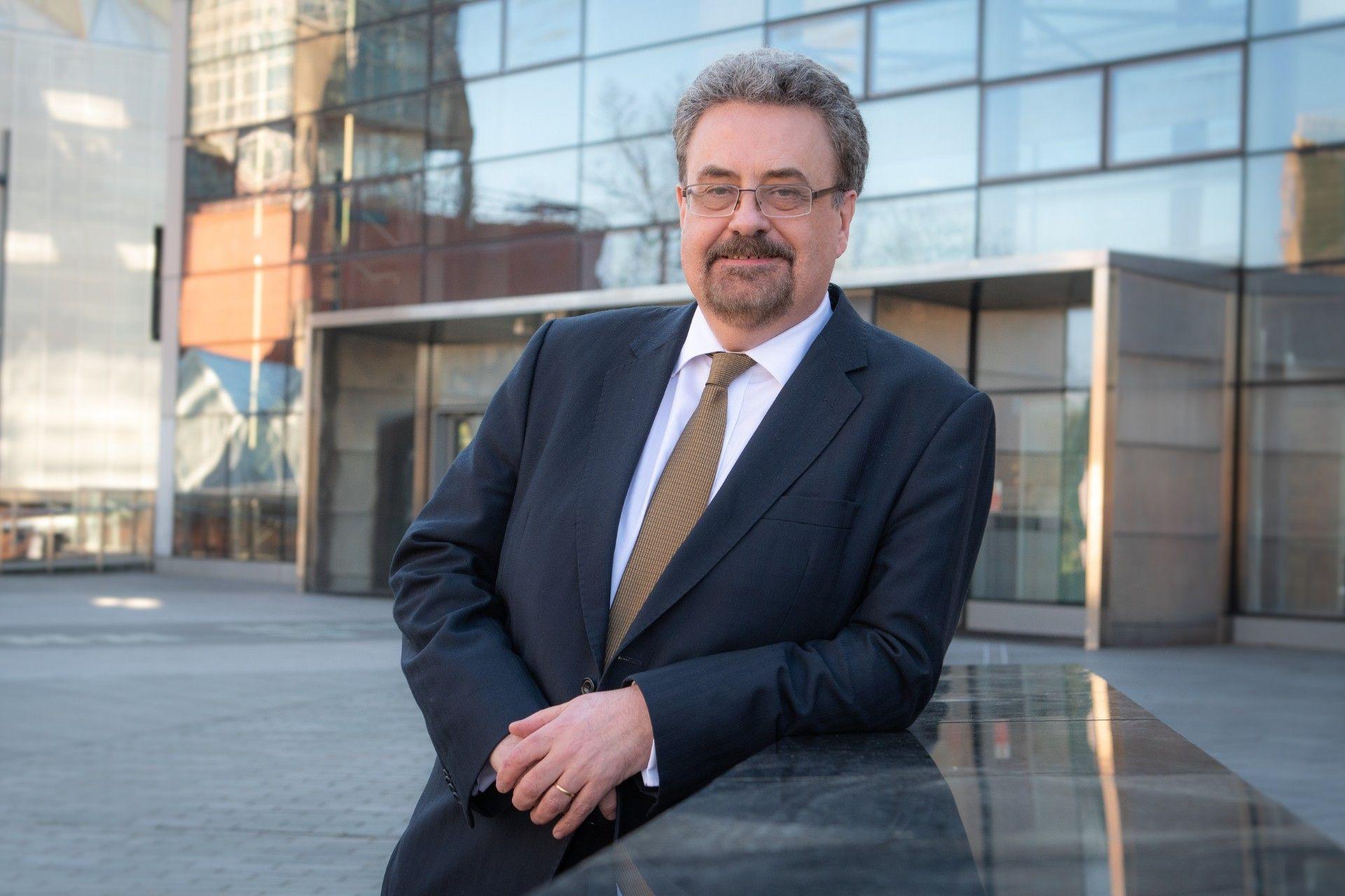 Prof Iain Gillespie in a blue suit and brown tie and wearing glasses, leans on a wall outside a university building