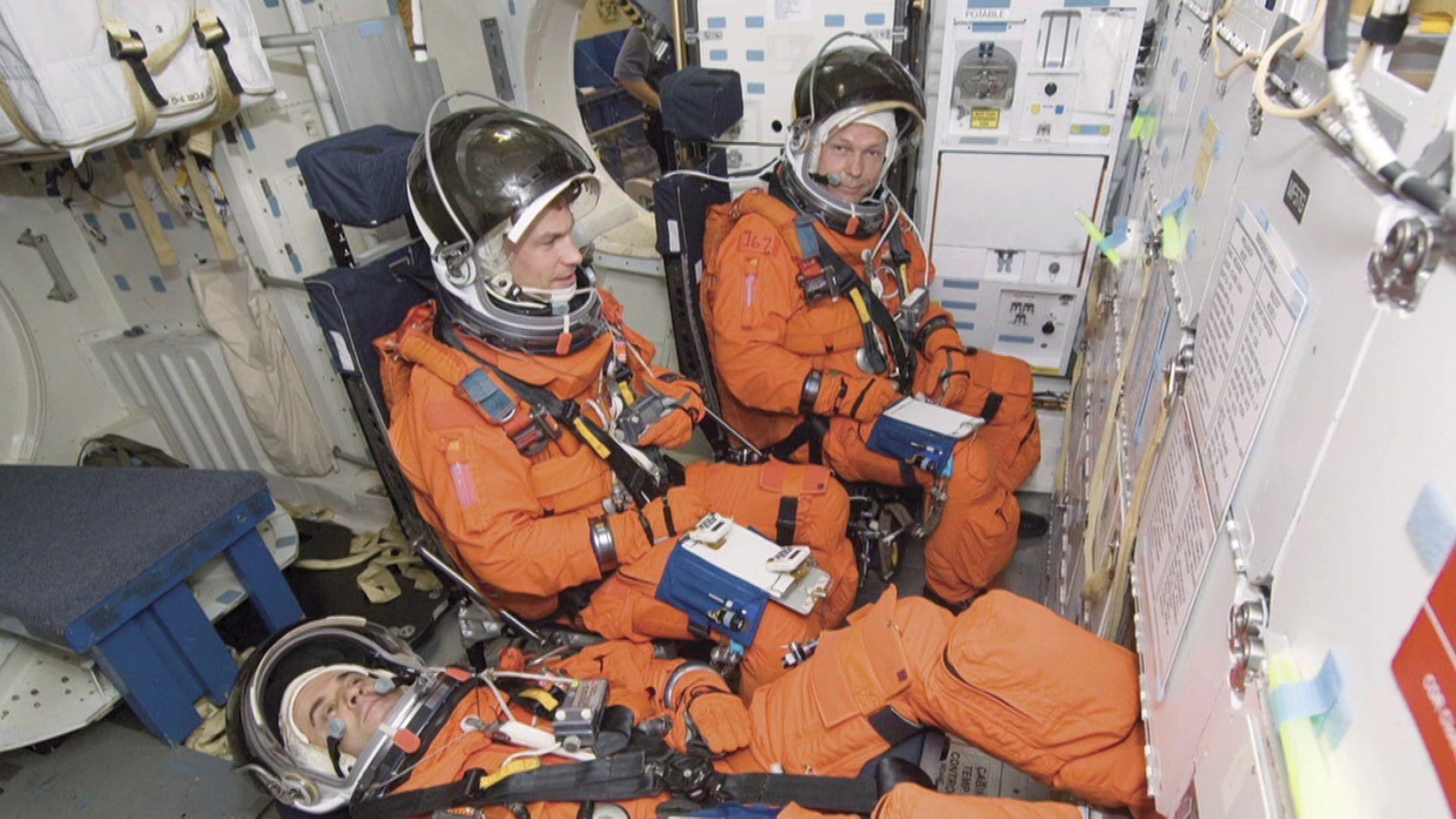 Three astronauts wearing orange space suits sit.
