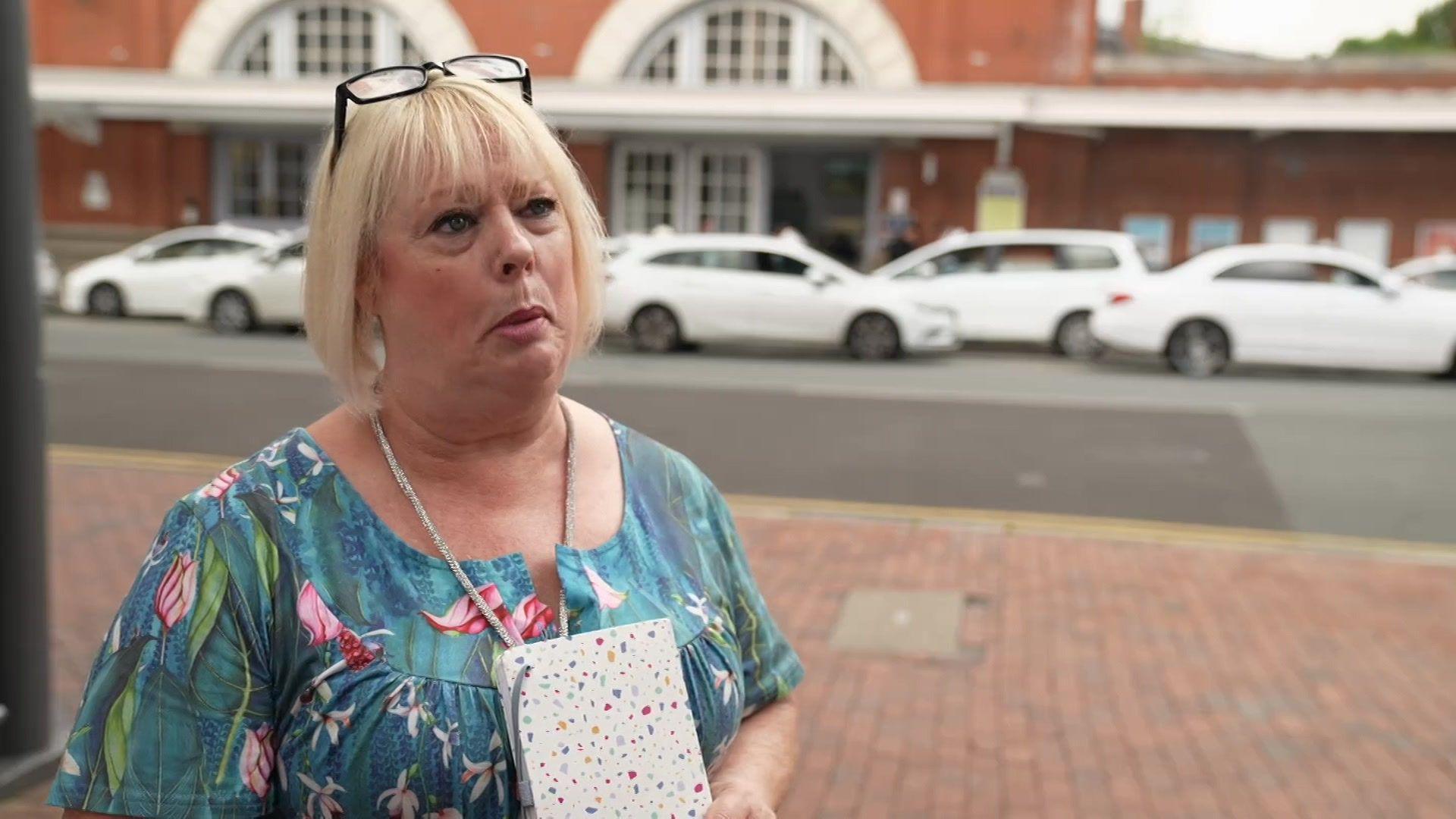 A woman standing outside a red brick train station with white taxis in the background.