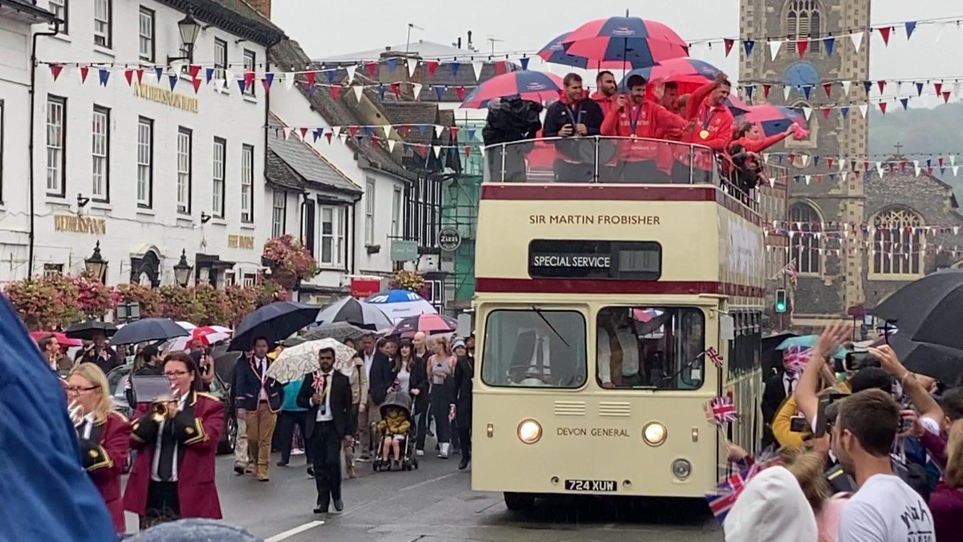 An open-topped bus making its way through Henley, with bunting hanged from shops from one side of the street to the other; a brass band making its way through the street, ahead of the bus; and on-lookers cheering the athletes, many of whom are holding umbrellas