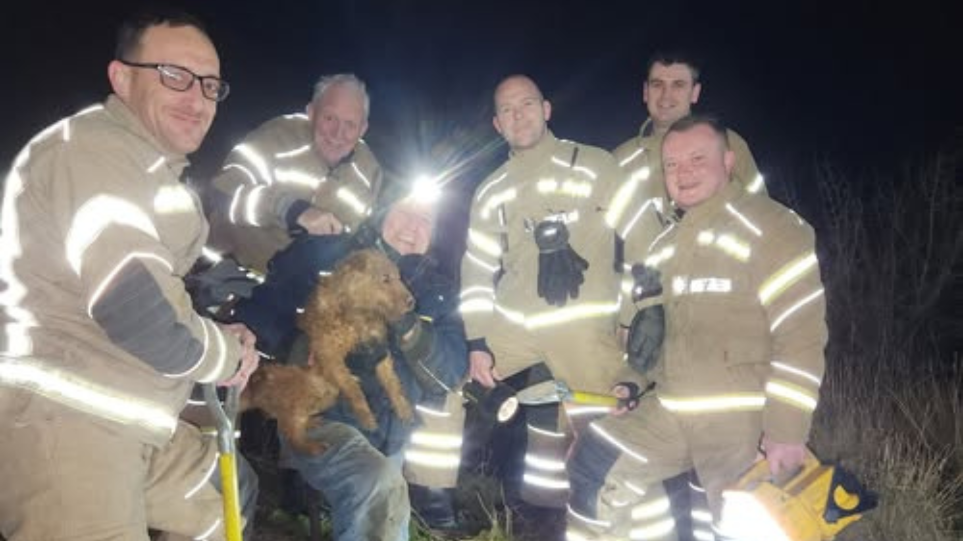 Five firefighters from Lincolnshire Fire and Rescue wearing their uniforms pose for a photo with terrier dog Branston and his owner David Benton. They are smiling at the camera after Branston was rescued.