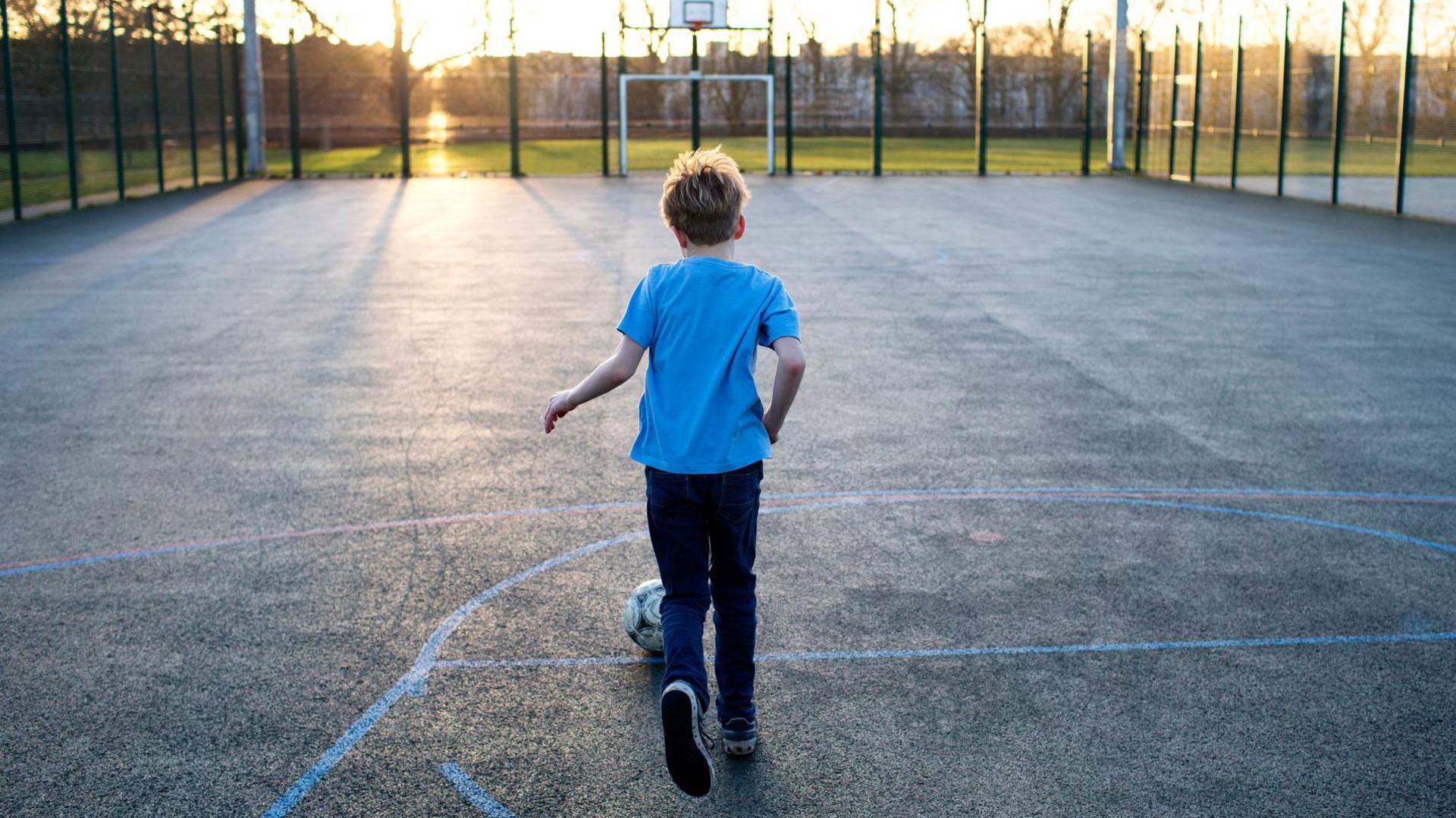 A thin boy wearing a blue T-shirt, dark trousers and trainers dribbles a football in an empty outdoor, fenced basketball/football court. The sun seems to be setting and there is a field outside the court area 