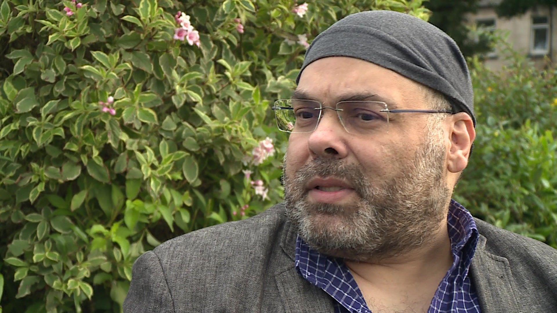 Dr Khalid Abdulraziq wears glasses, a bandana on his head and a blue and white checked shirt with a grey jacket. Green foliage is visible behind him.