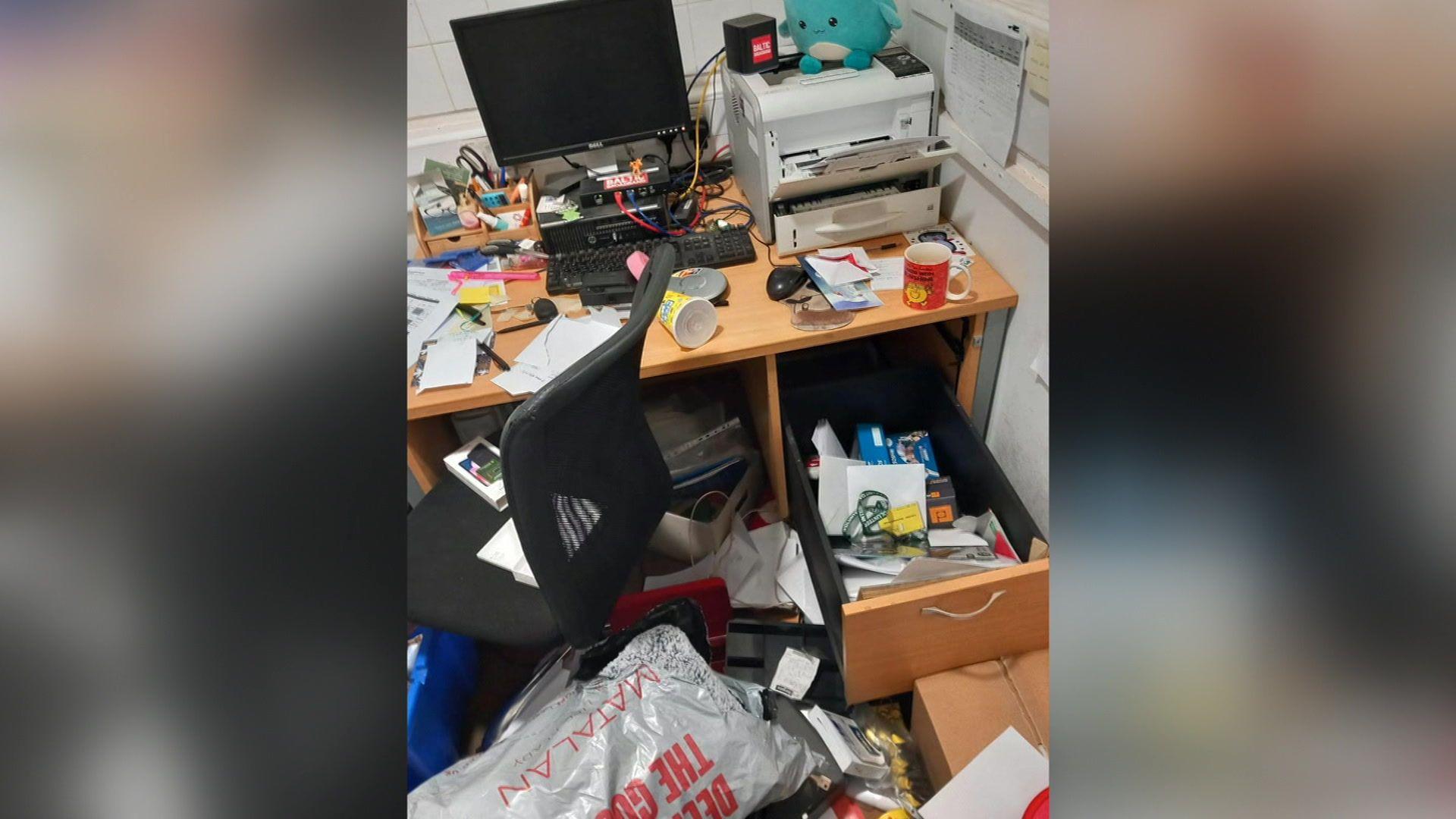 A light brown wooden desk, with a computer monitor and printer, is covered in mess. A drawer has been left open.