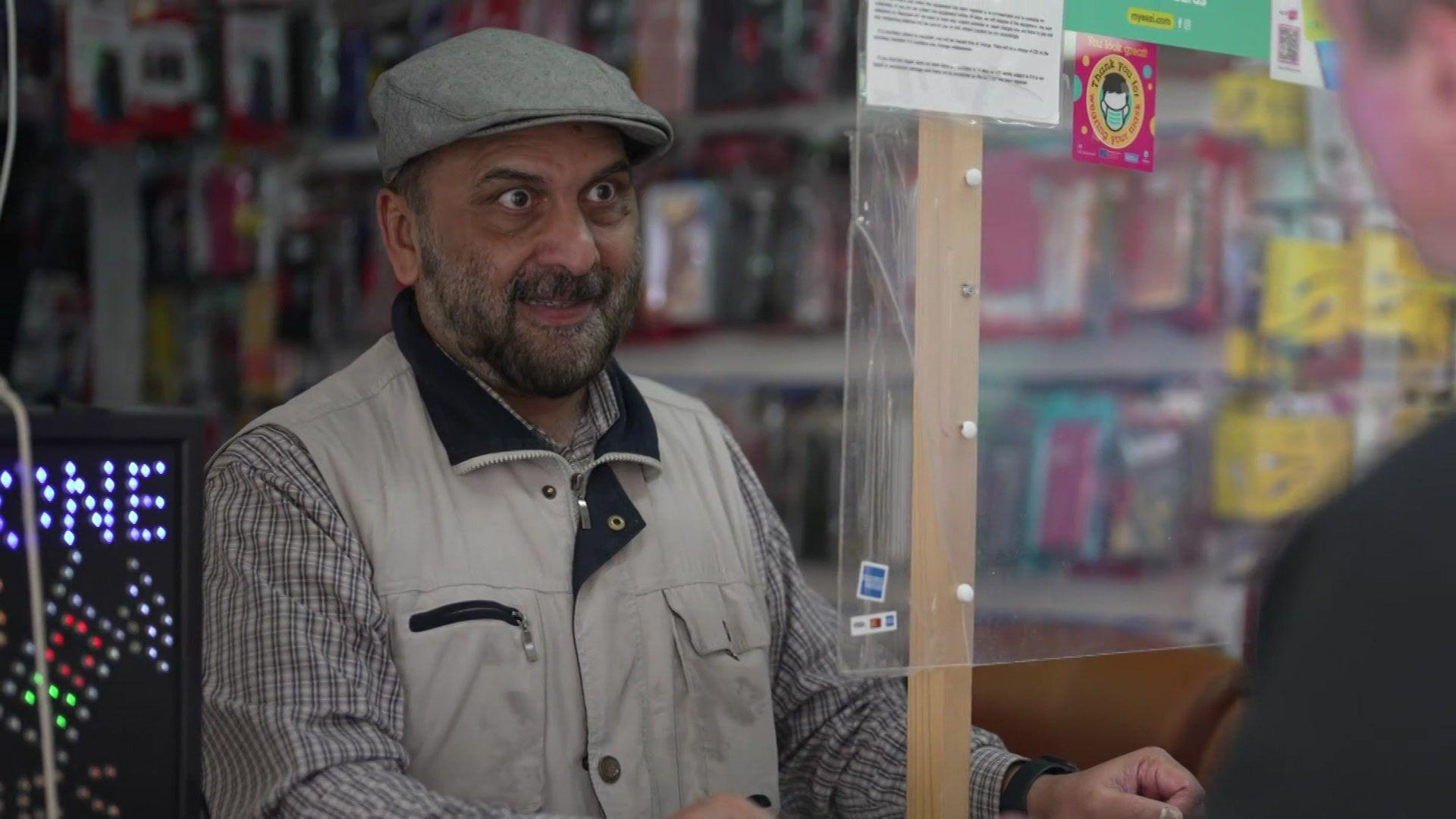 Hussein wears a grey baker boy cap, beige workman gilet and a black and beige check skirt. He stands inside his mobile shop.