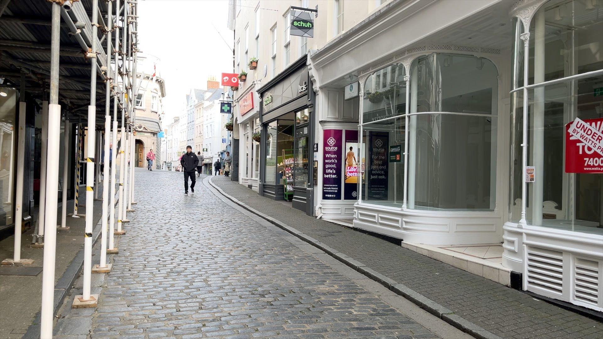 A cobbled street with shops either side and scaffolding outside the buildings on the left side with a few people walking along the street