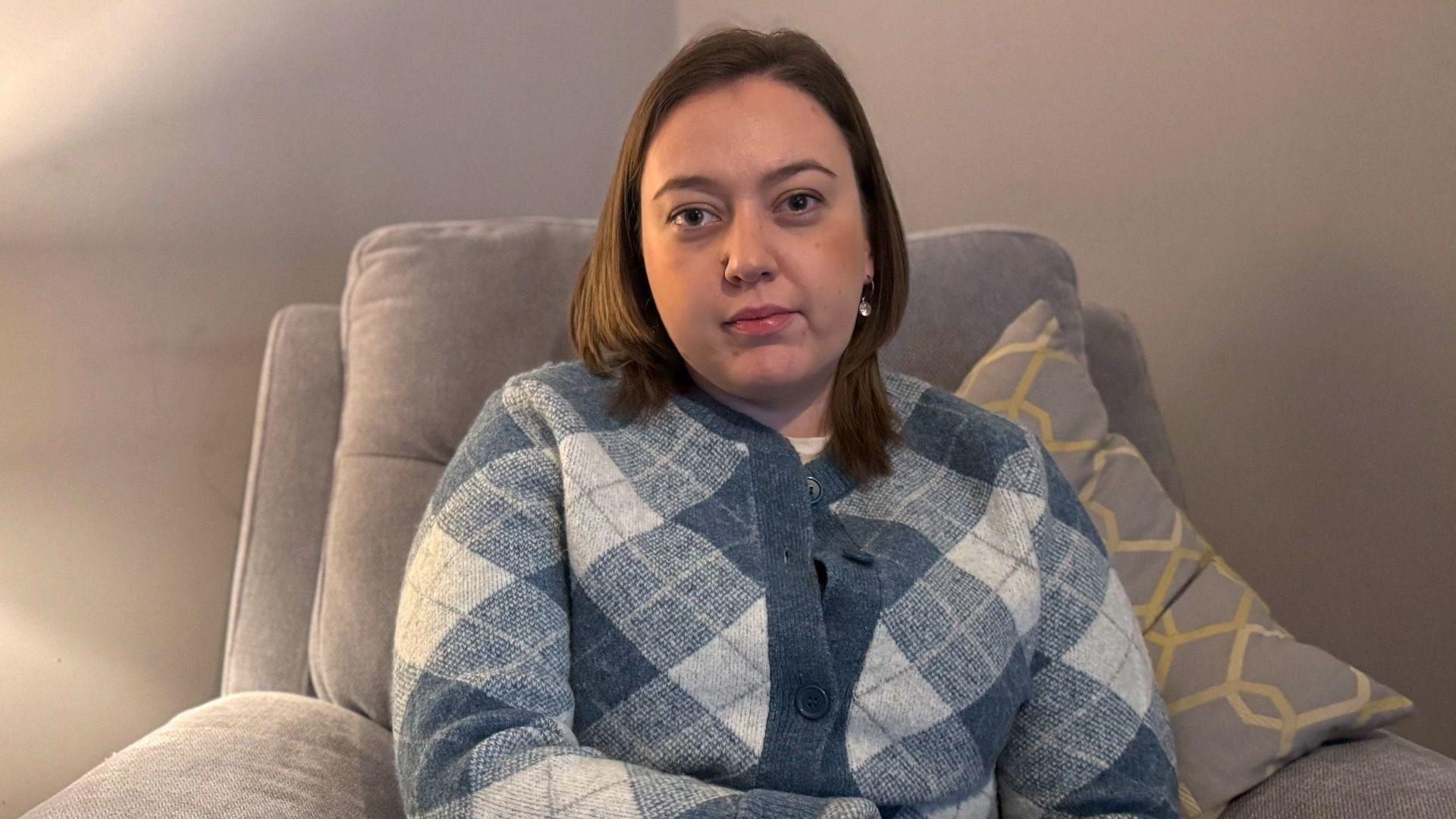 A woman with shoulder length brown hair looks toward the camera. She is wearing a blue and cream patterned cardigan and is sitting on a grey chair. 