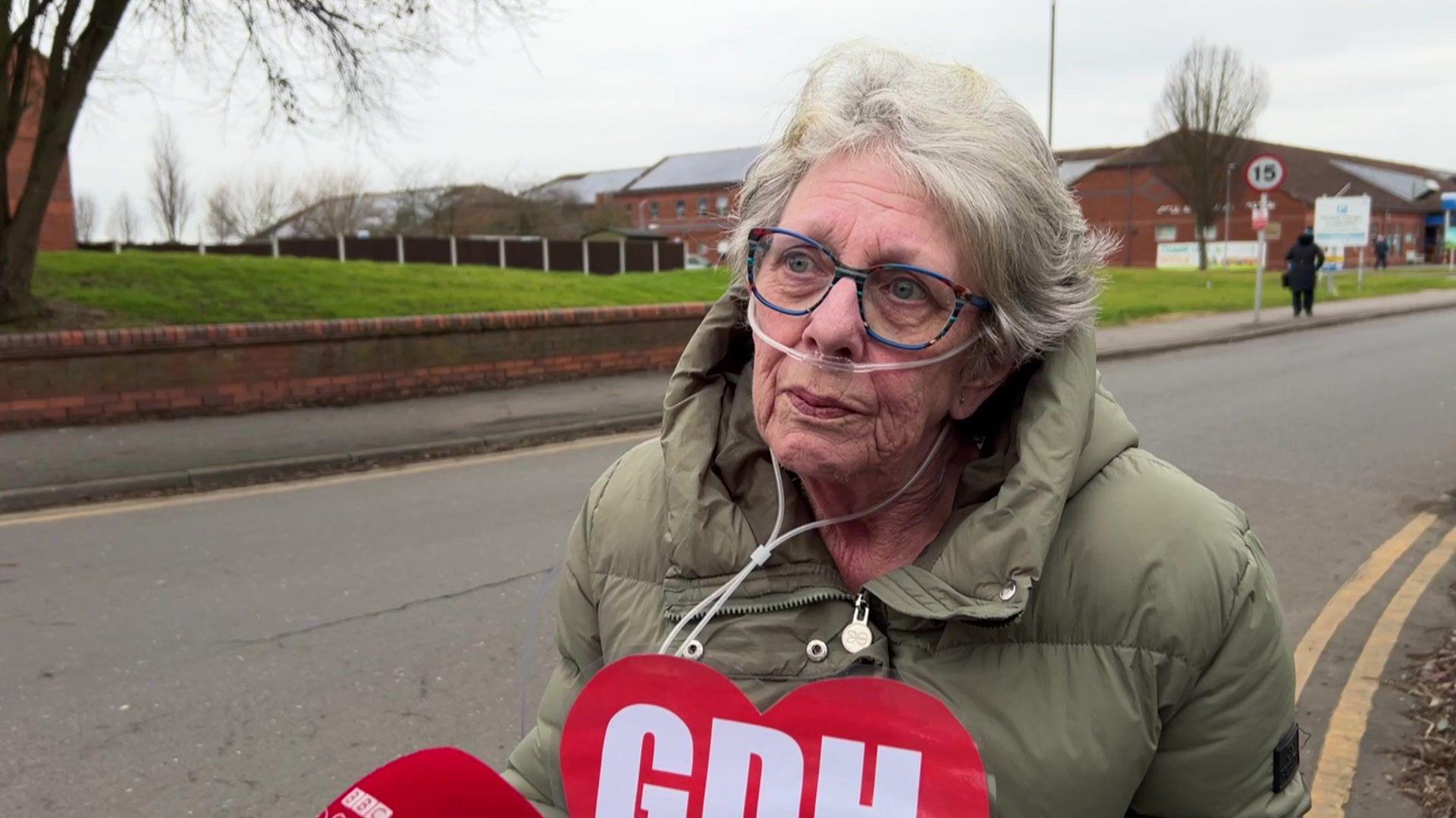 A head and shoulders shot of Jean Thompson - a woman with short grey hair, glasses and a nasal cannula for oxygen. She is sitting in a wheelchair, which is not visible in the photo, on a road near a brick building. She is wearing a green puffer jacket and is holding a red heart-shaped sign with letters on it. The sky is overcast.