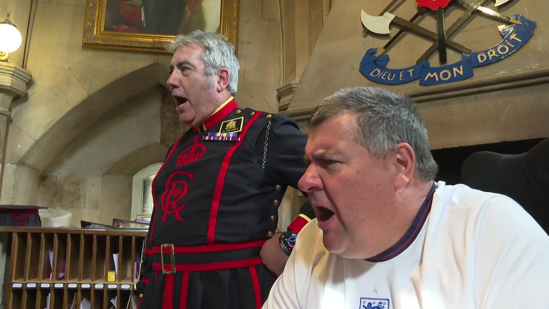 The two men in the historic room watch the game and react with a shout. A fireplace with a crest bearing the words “Dieu et Mon Droit” is visible behind them.