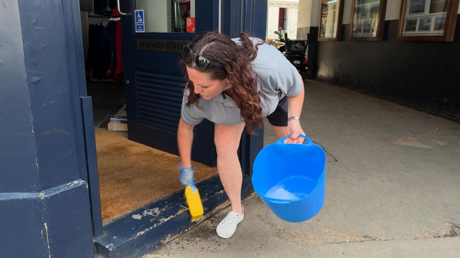A woman in a grey T-shirt holding a blue bucket of water and scrubbing brush cleaning the entrance to a shop