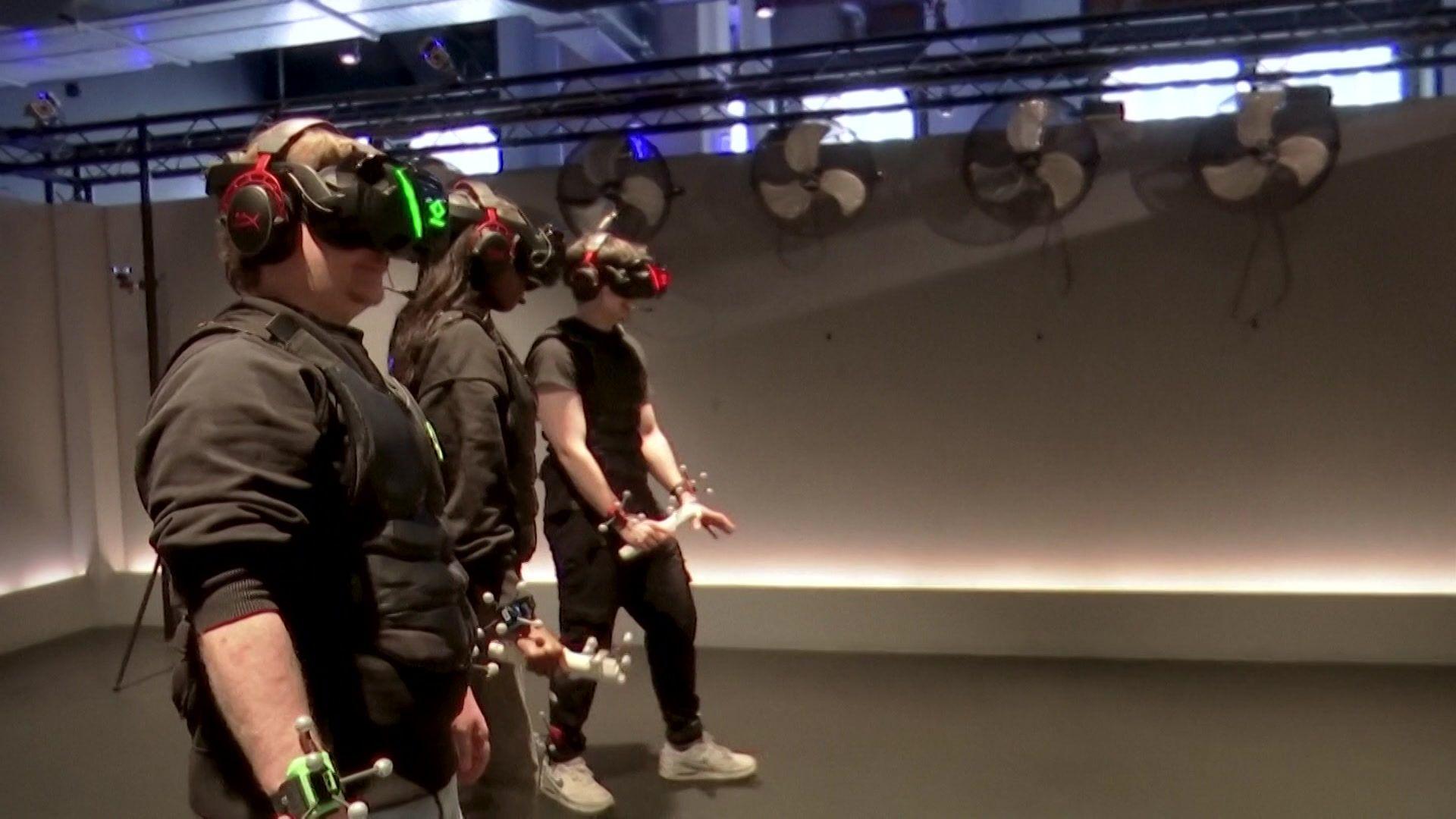 Three people stand in a line in a large grey room with virtual reality headsets on over their eyes. They are all dressed in black with contraptions on their wrists with sticks and balls on. 