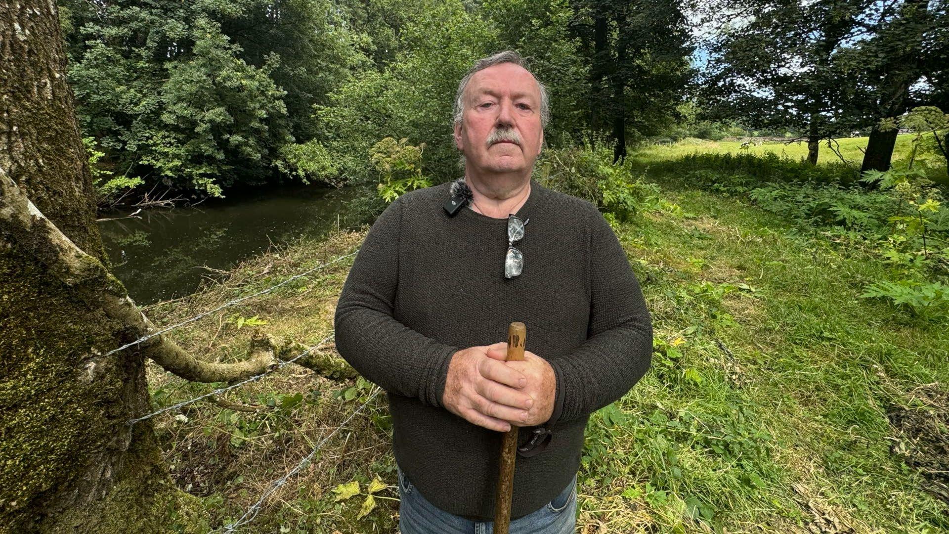 A man with short grey hair, a moustache, glasses around his neck and a brown jumper. He holds a brown wooden stick in his hand  