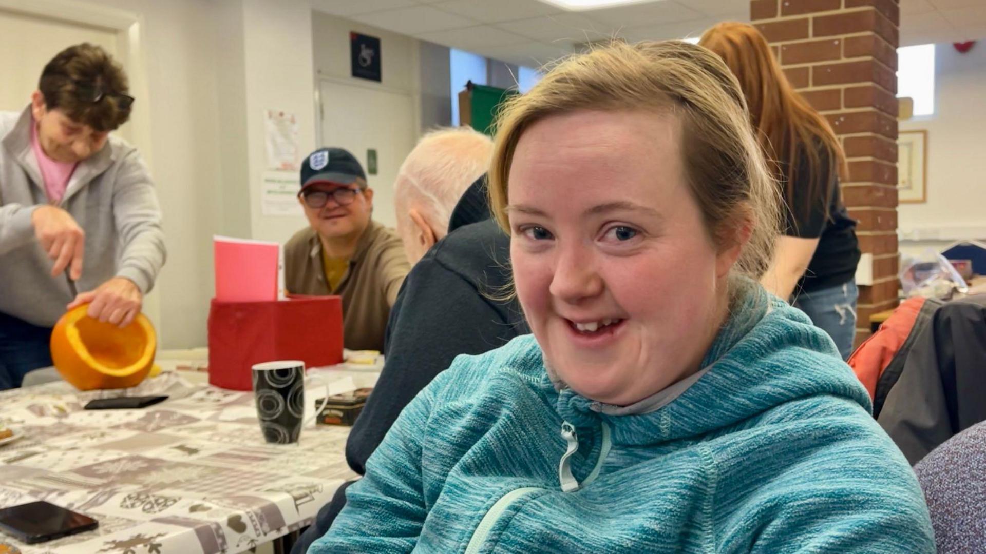 A lady is sitting and smiling dressed in a green hoodie. Behind her is a table with a cream patterned table cloth with four people sat and standing around it. One lady dressed in a grey fleece is carving a pumpkin.