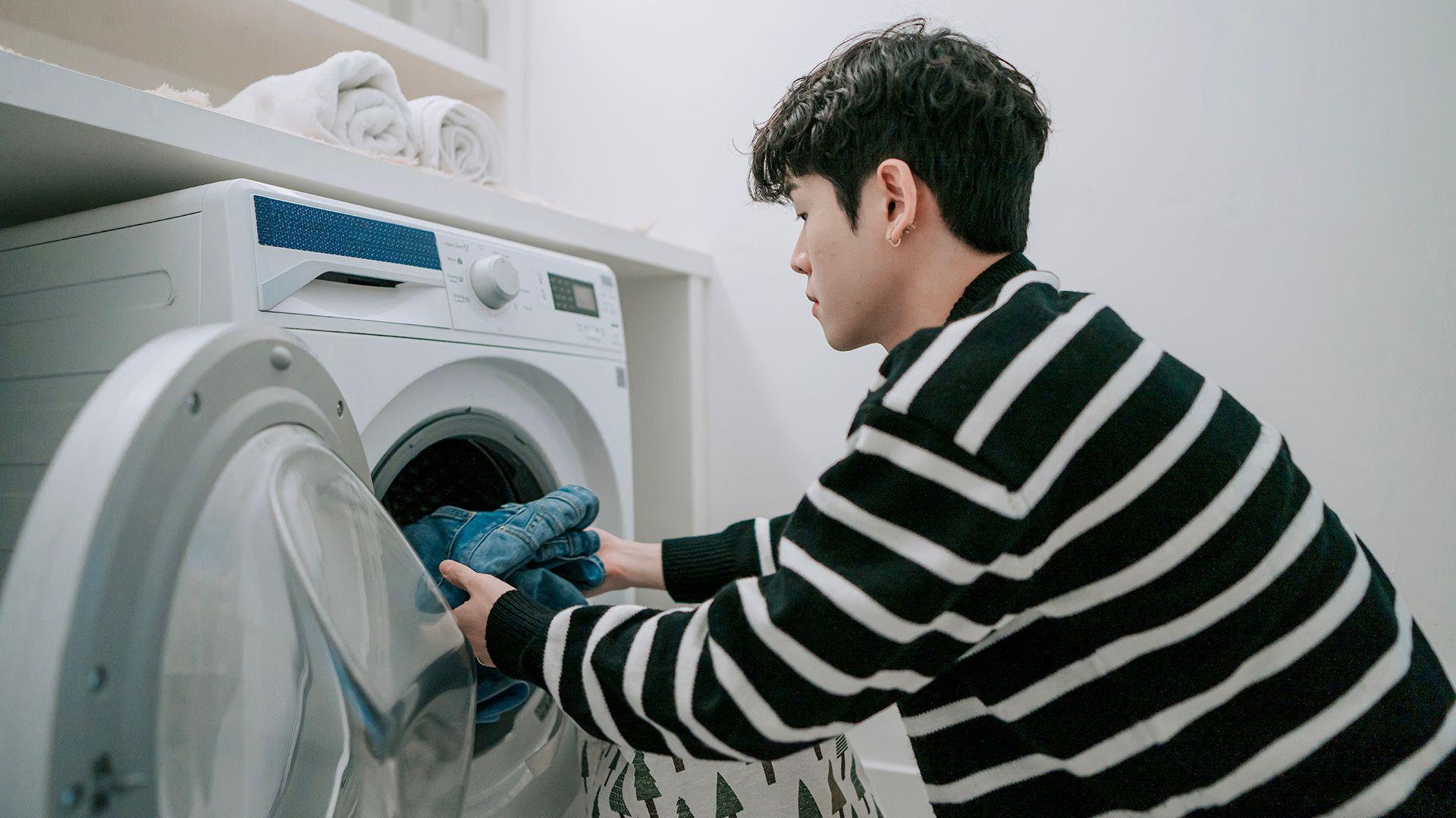 Stock photo shows a person wearing a black and white striped top loads clothes into a washing machine in a laundry room at home.