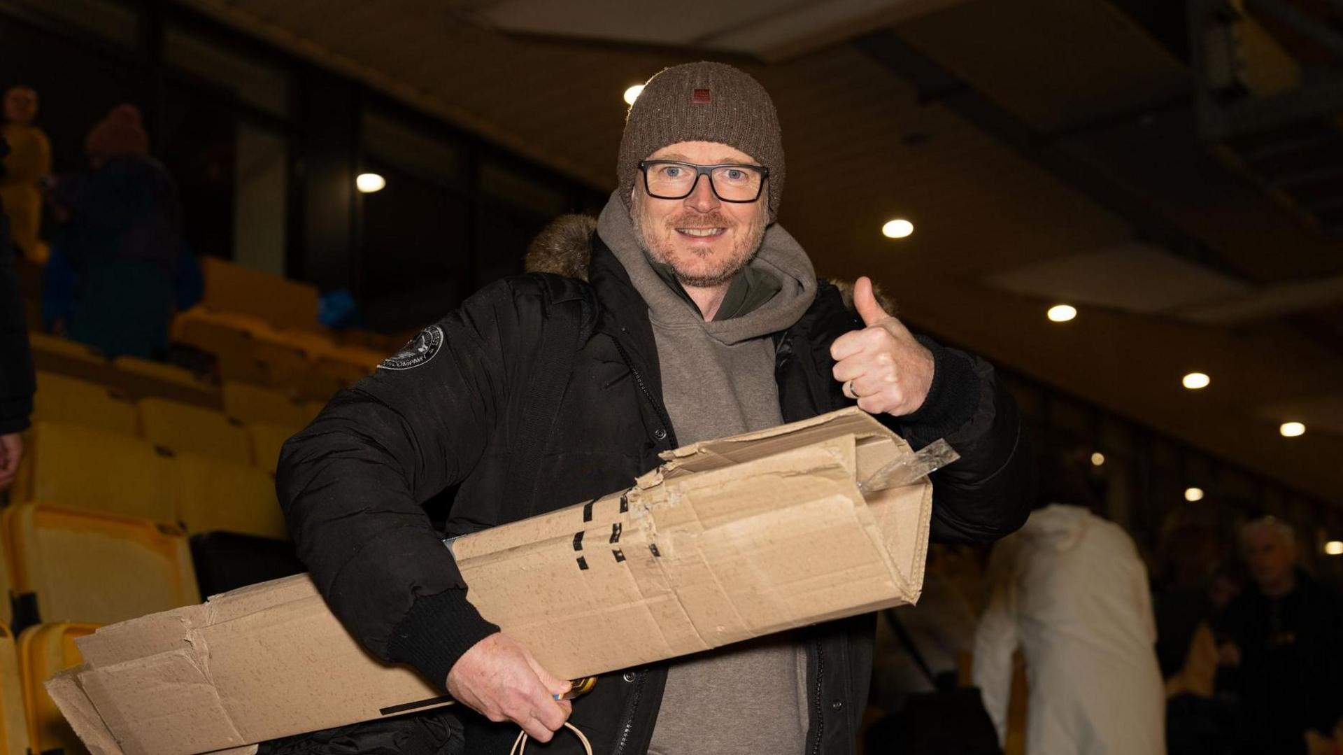 A man in a hoodie, dark puffa coat and woolly hat gives a thumbs up sign as he carries cardboard under his arm in a stadium stand with yellow seats. It is dark but the stand is lit.