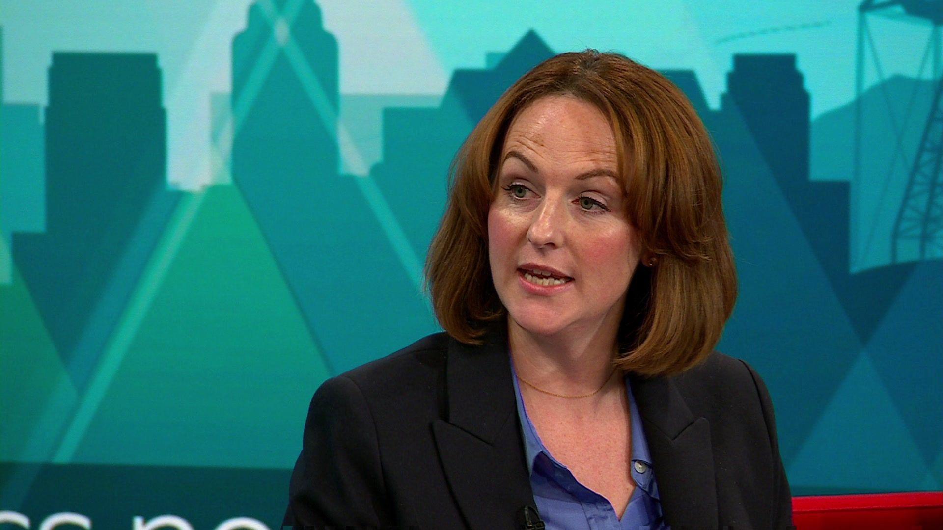 Lola McEvoy is pictured mid sentence sat on the BBC Politics North red sofa with the studio's teal-coloured cityscape graphical backdrop behind her.