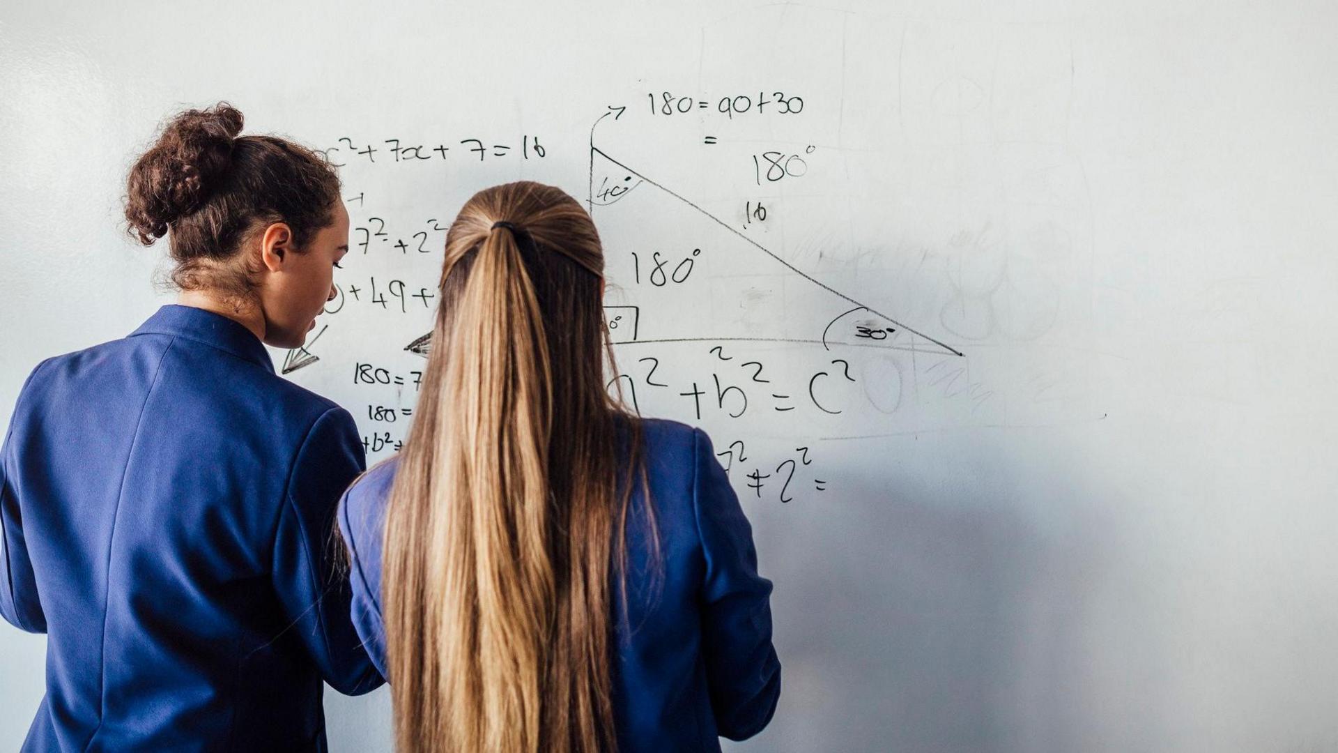 The picture shows two schoolgirls in blue uniforms standing in front of a whiteboard on which is written  a mathematical equation 