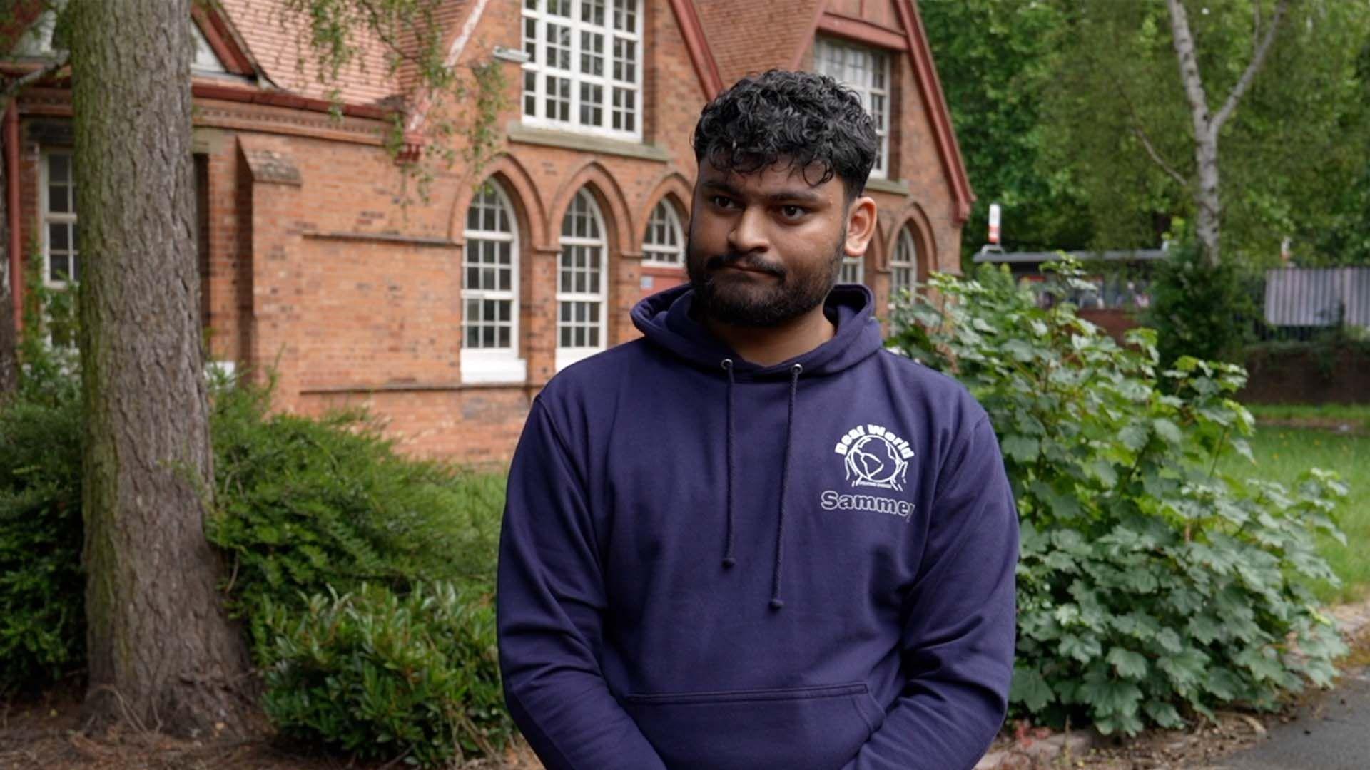 Sammey Ahmed is wearing a dark blue jumper with his name on, he's standing in front of a brick building with a tree next to him. 