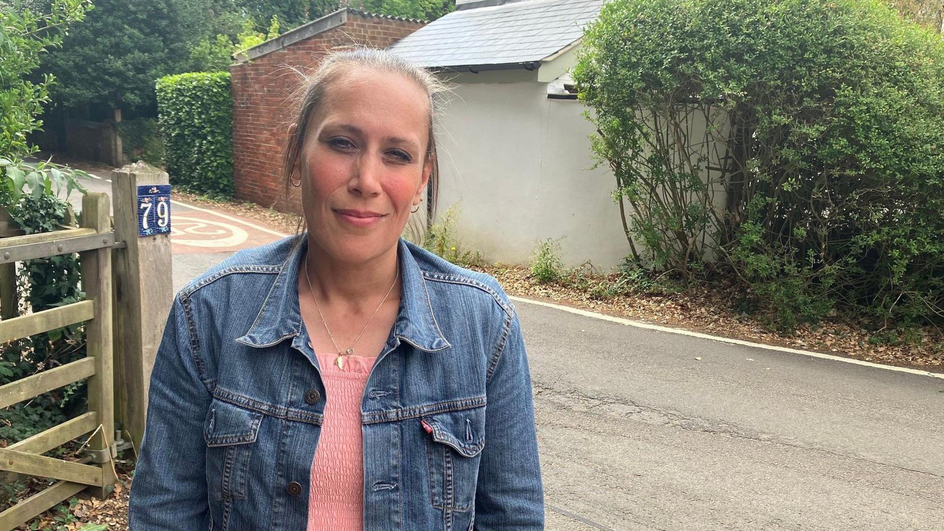 A picture of a woman stood by the side of Walton Road in Wavendon village near Milton Keynes. She has a pink top on, under a blue denim jacket and her hair has been pulled back. There is a gate to her right.