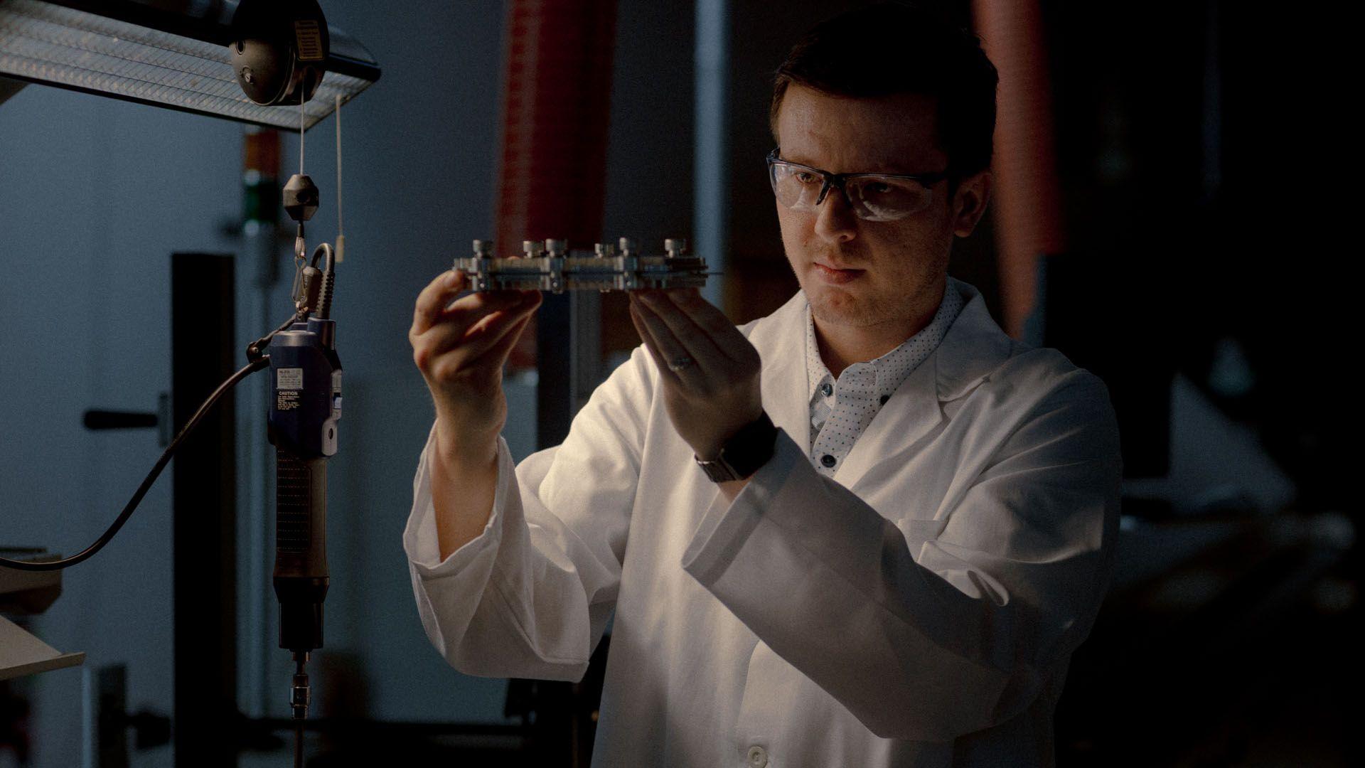 A man standing holding a component wearing a white lab coat and safety goggles 