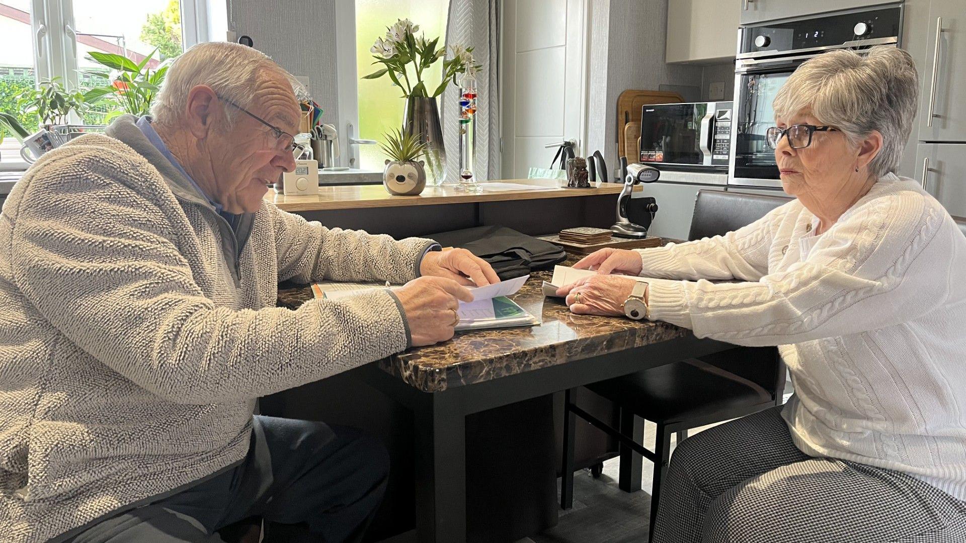 An elderly man and a woman in her 60s sit opposite each other at a marble top kitchen table. They both have folders of paperwork in front of them and they are looking towards each other. Both are wearing glasses. He has white hair and she has grey hair. He is wearing a beige fleece and blue trousers, she is wearing black and white check trousers and a white arran jumper, as well as a large white-faced wristwatch. A window, vase of flowers, microwave and built-in oven are visible behind them. 