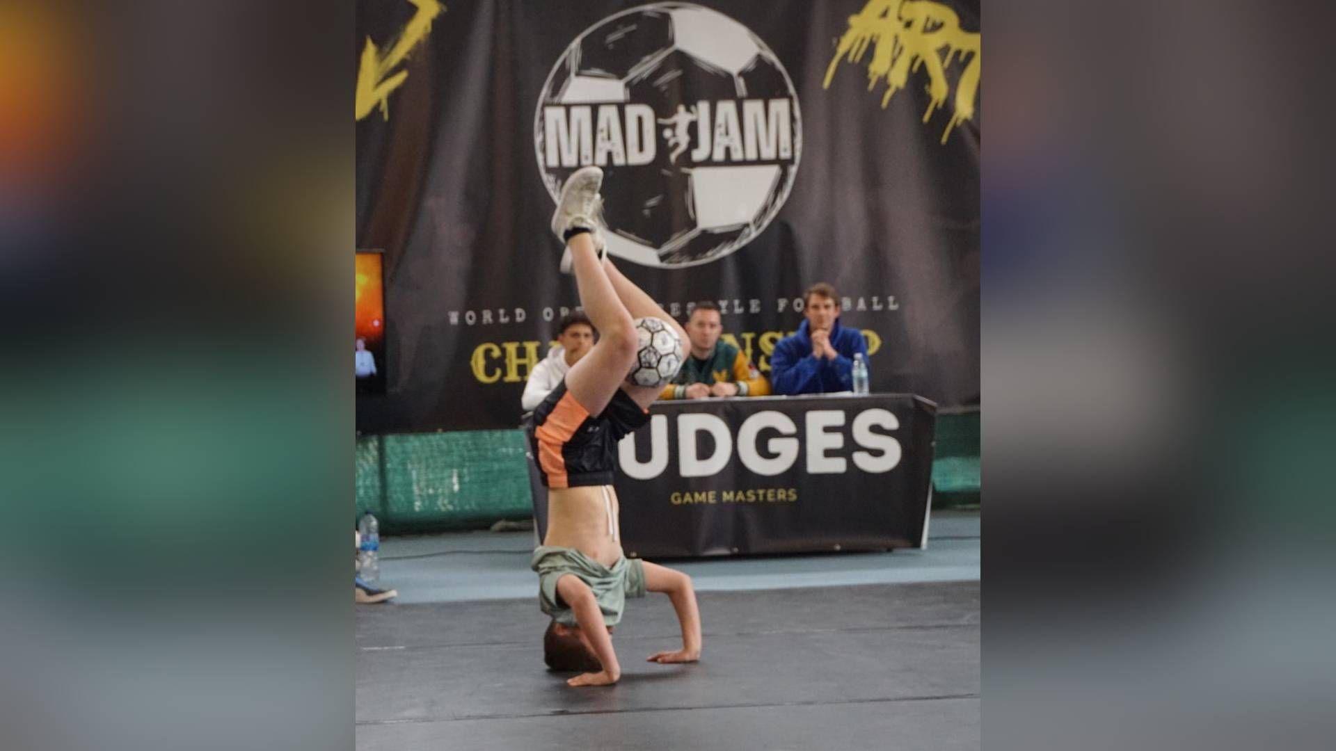 James doing a handstand with a football between his knees. A panel of judges sit on a table behind him, with a black curtain behind them which says Mad Jam inside a football graphic.