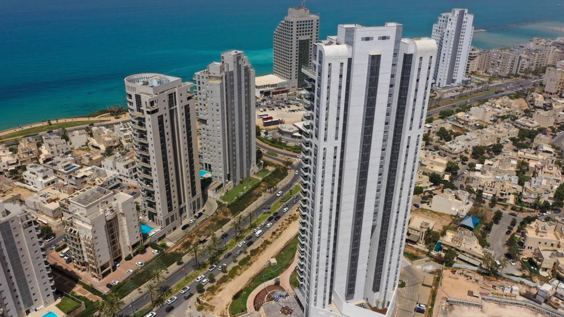 A birds eye view shot shows skyscrapers near the coast. The water is blue and there is a large building in the foreground with other tall buildings and houses behind. 