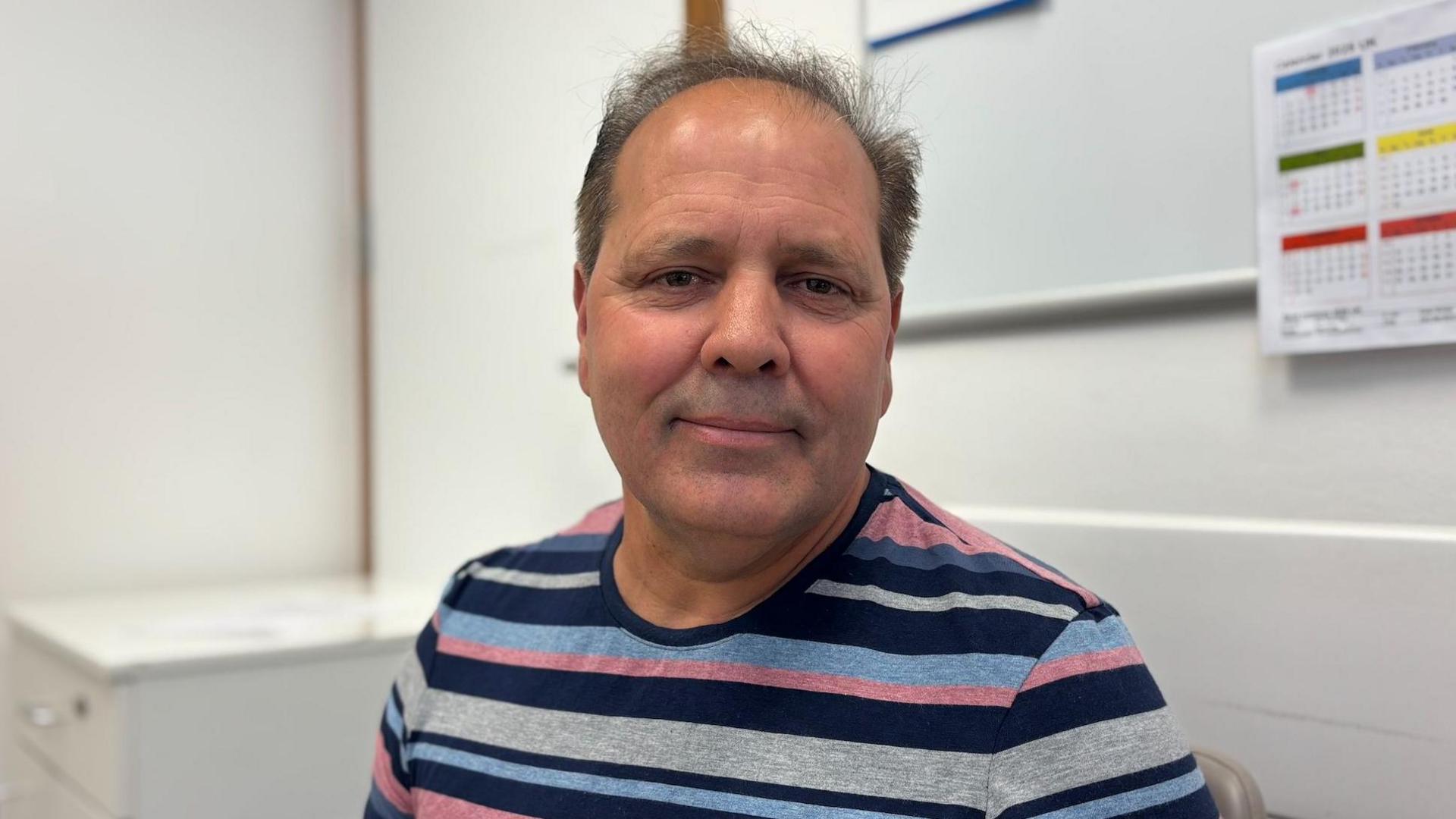 Head shot of Albert Jardim wearing a pink blue and grey stripe t-shirt. He is sat down in a clinical office.